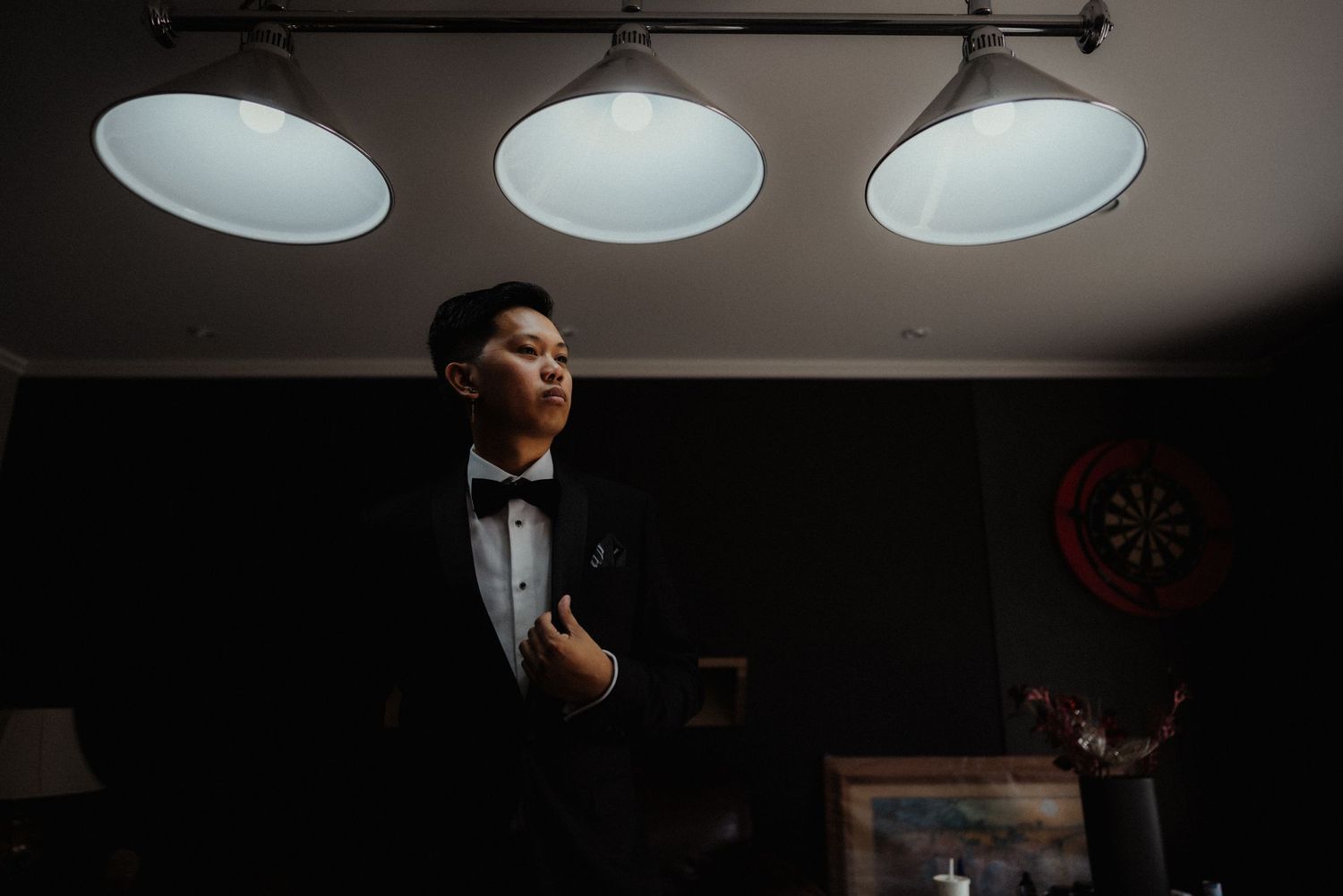 Groom in a black tuxedo and bow tie stands in a dimly lit room beneath three pendant lamps.