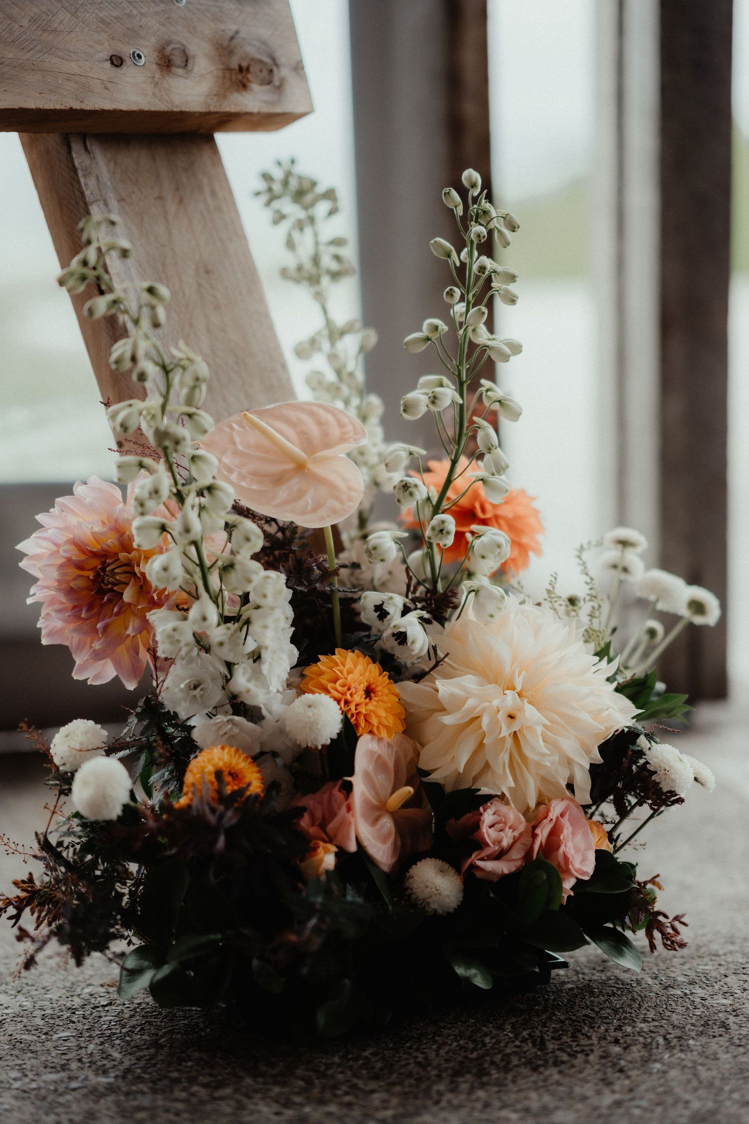 Wedding floral arrangement of dahlias, orange blooms, anthurium and white flowers sits on the floor beside a wooden easel.