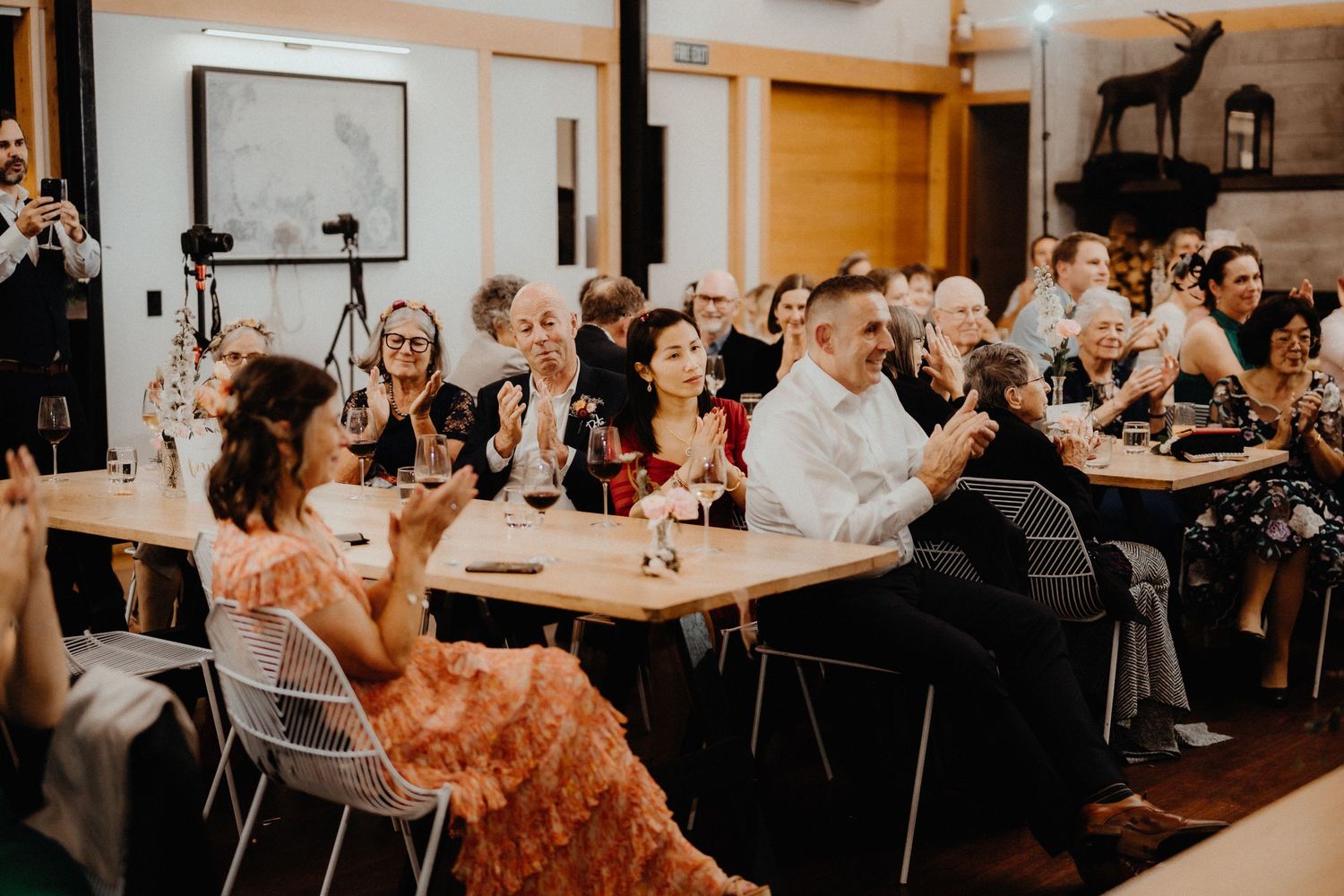 Wedding guests seated at long wooden tables smile and applaud during a reception in a rustic timber venue.
