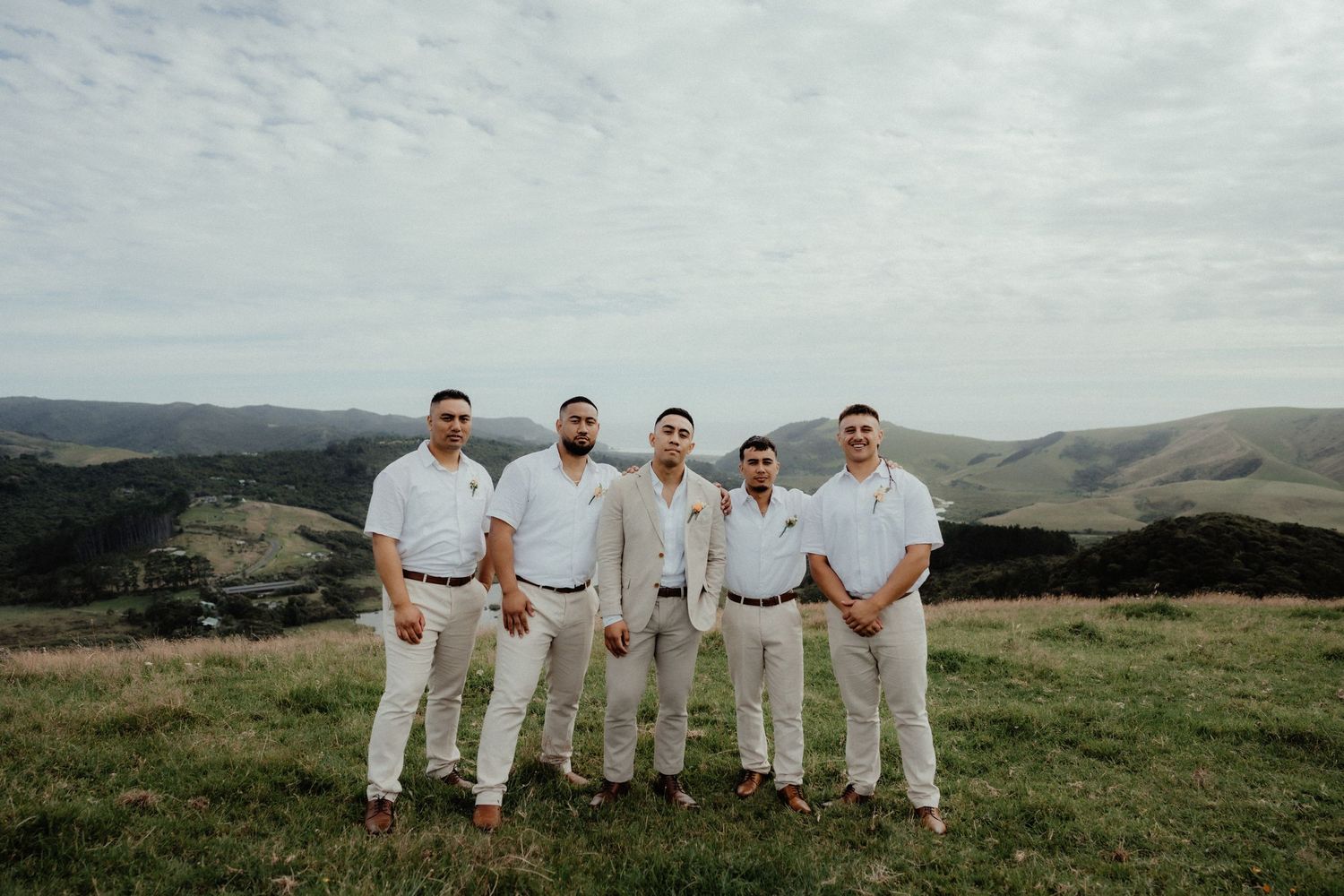 Five groomsmen in white shirts and neutral trousers stand together on a grassy hillside with rolling green hills behind them.