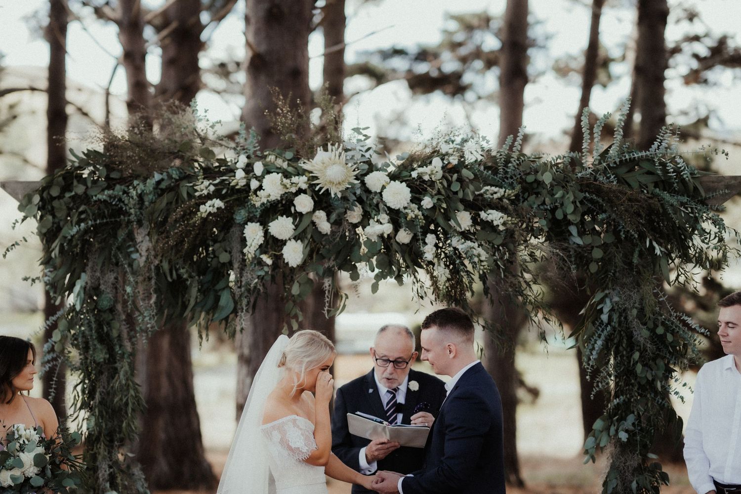 Bride wipes a tear as the groom holds her hand during their outdoor wedding ceremony beneath a lush floral arch.