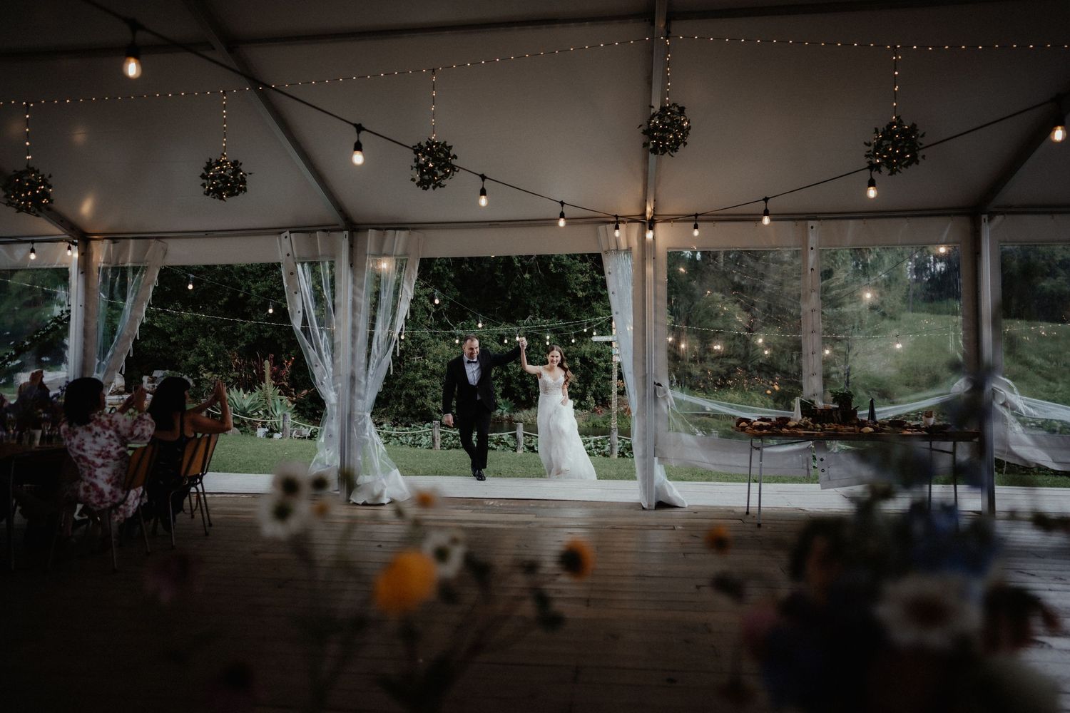 Bride and groom walk hand in hand toward a decorated marquee with draped curtains and string lights at their wedding reception.