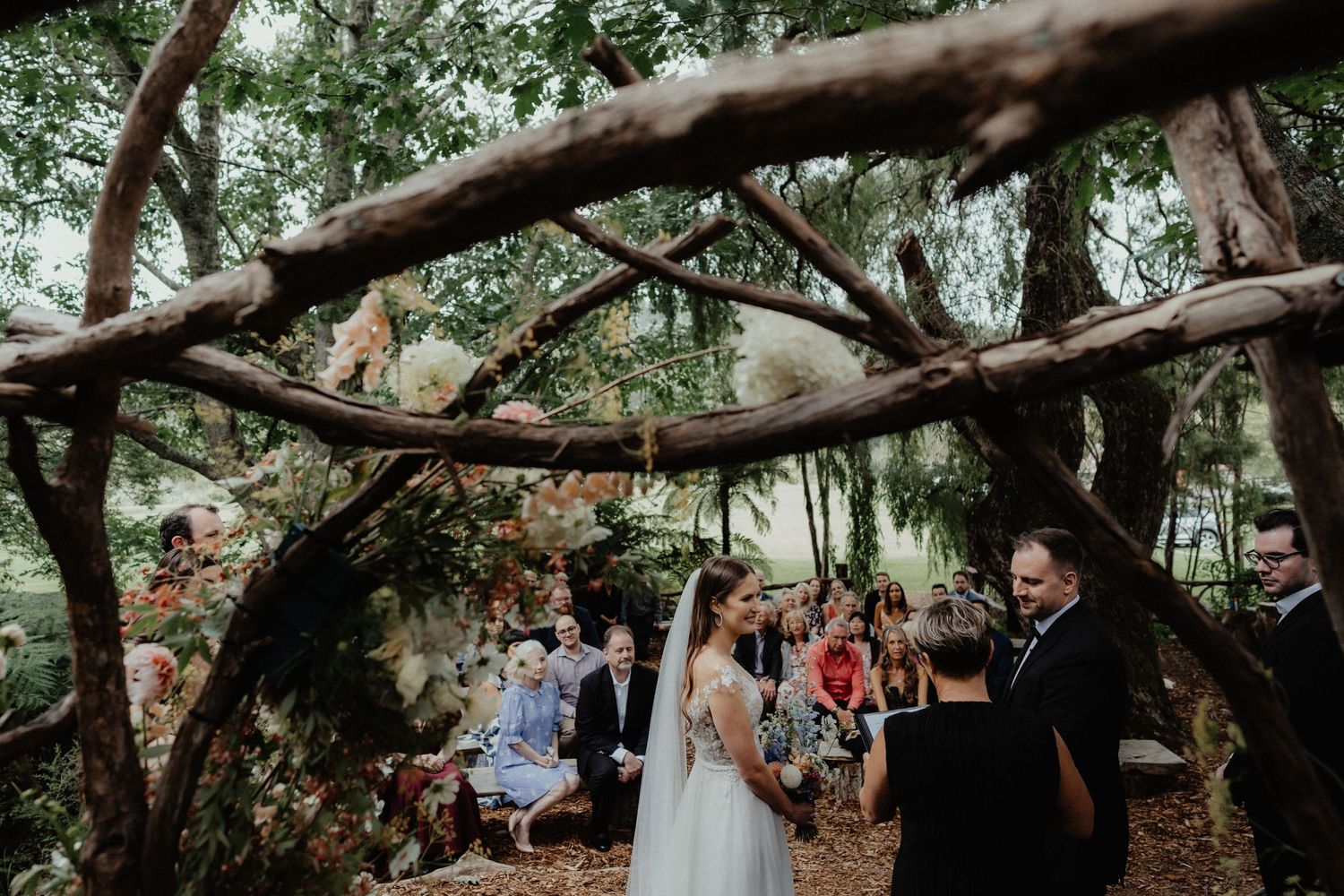 Wedding guests sit under a canopy of rustic tree branches as the bride and groom stand during an outdoor ceremony.