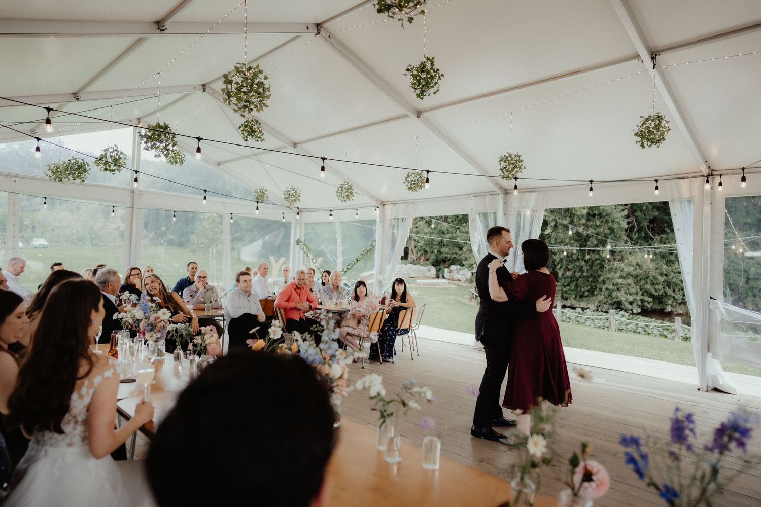 Bride and groom share a dance in a decorated wedding marquee as seated guests watch from reception tables.