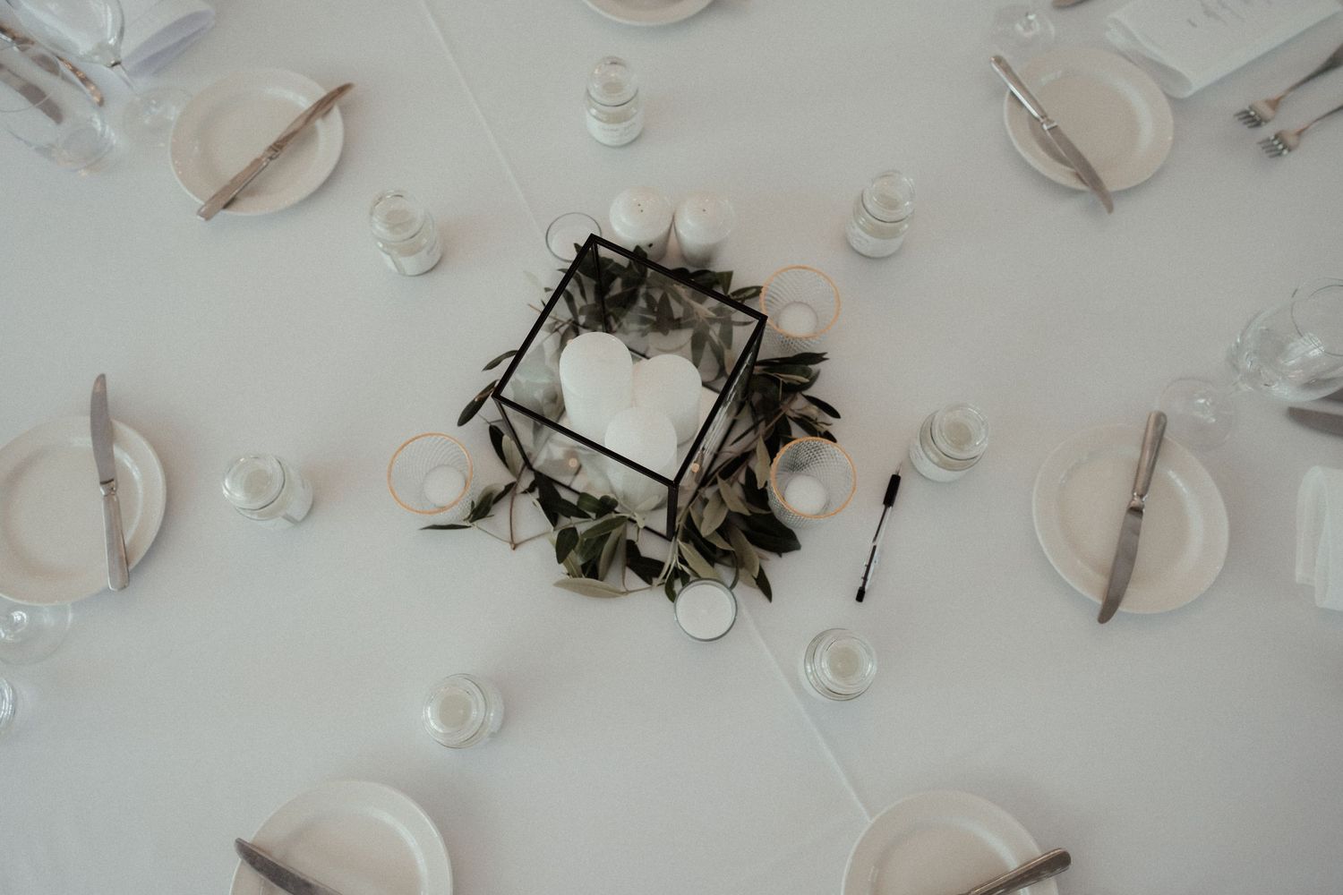 Overhead view of an elegant wedding reception table with white plates, glassware, and a greenery-wrapped glass centerpiece holding a heart.