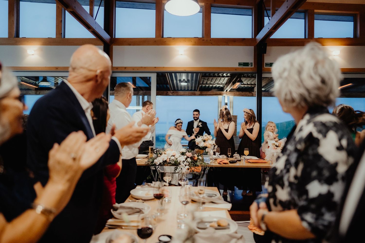 Wedding guests applaud and cheer during speeches at a reception in a bright venue with wooden beams.