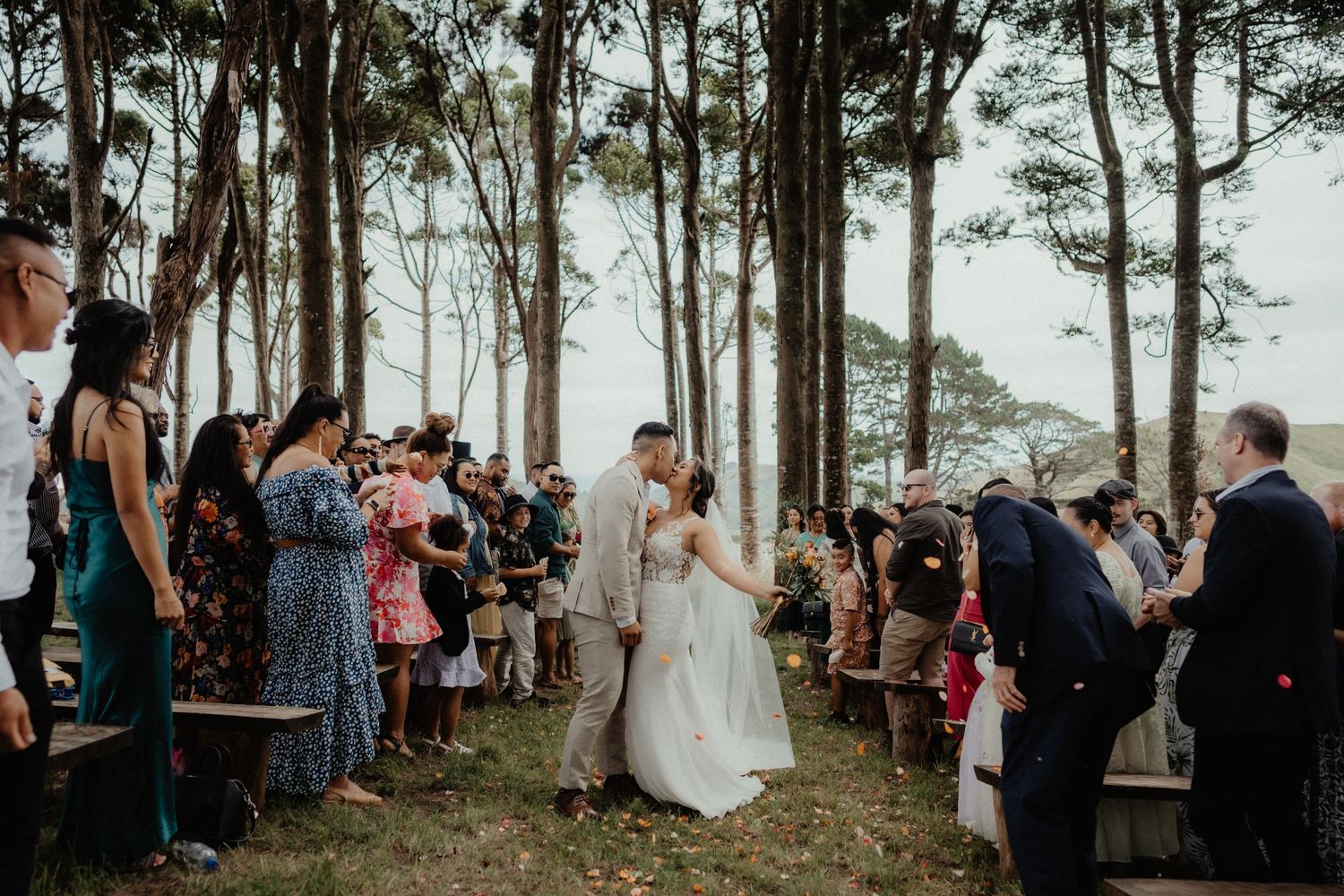 Newlyweds share a kiss in a forest aisle while seated guests applaud and cheer at their outdoor wedding ceremony.