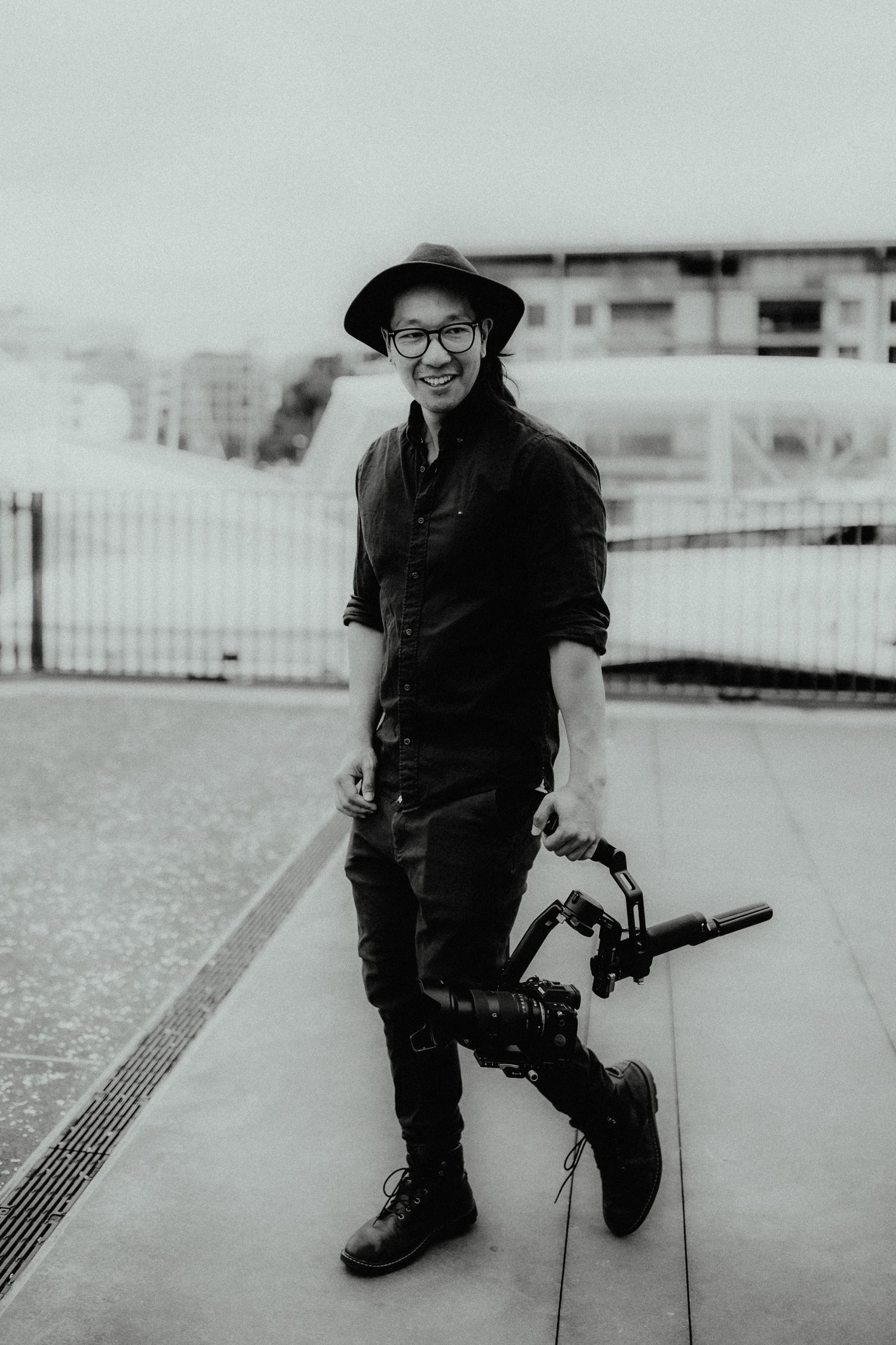 Black and white photo of a smiling man in a wide-brimmed hat and glasses holding a camera gimbal stabilizer on a pavement.