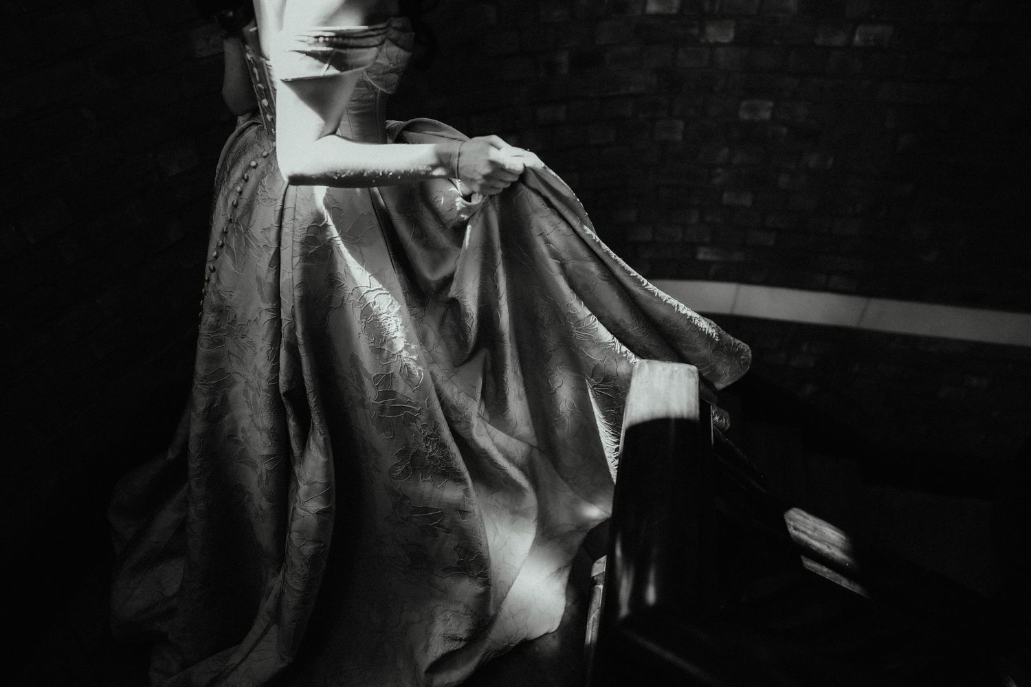 Black and white photo of a woman in a flowing satin gown gathering her skirt while standing beside a dark staircase.