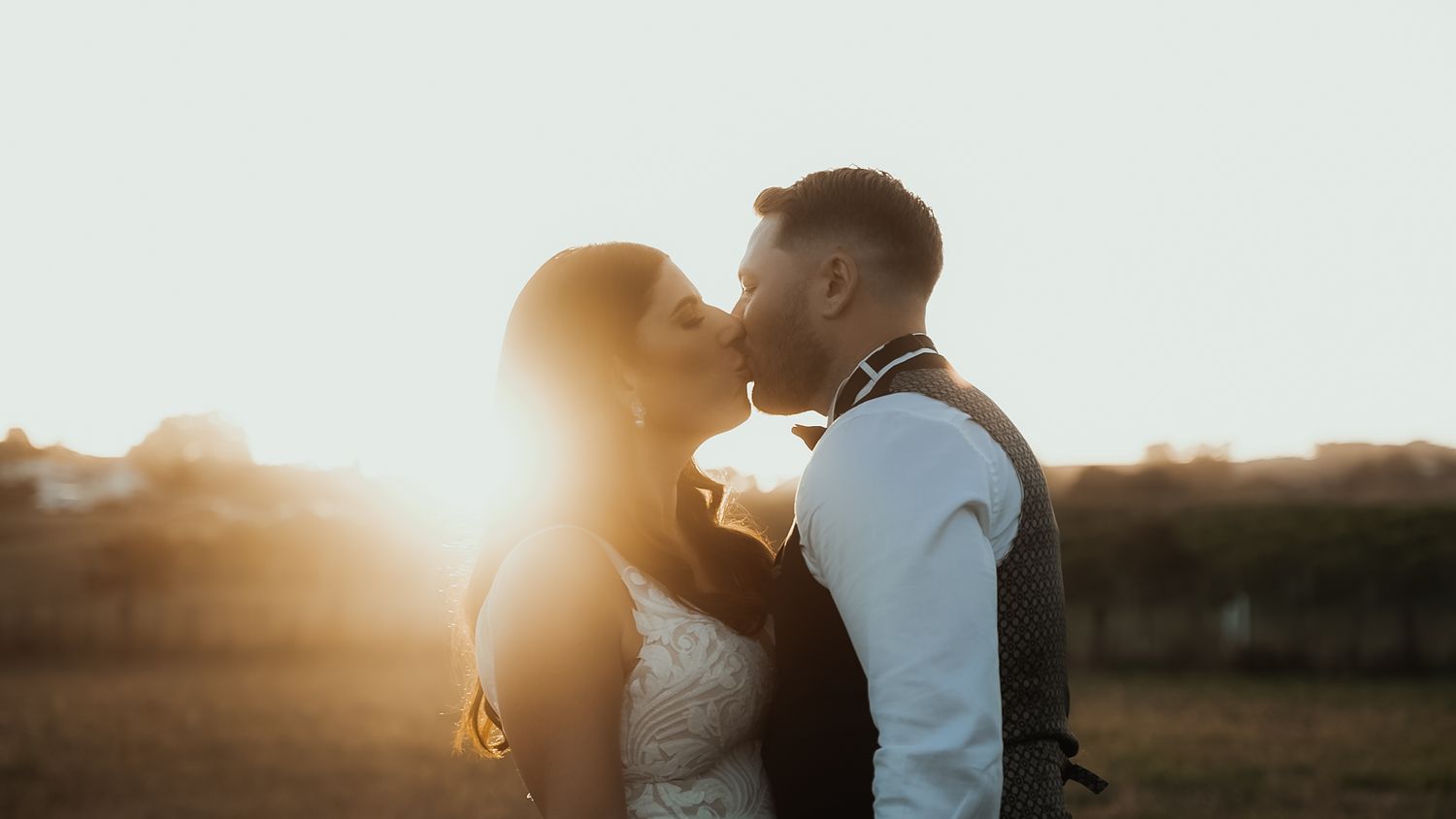 Bride and groom share a romantic kiss in warm golden sunset light with a glowing sun flare behind them.