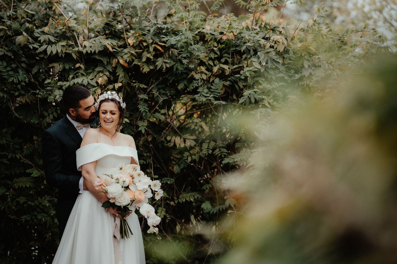 Groom embraces his smiling bride in a white off-the-shoulder gown holding a bouquet in front of lush green foliage.