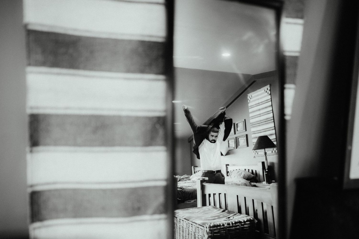 Black and white photo of a groom stretching his arms overhead in a bedroom, seen through a mirror reflection.