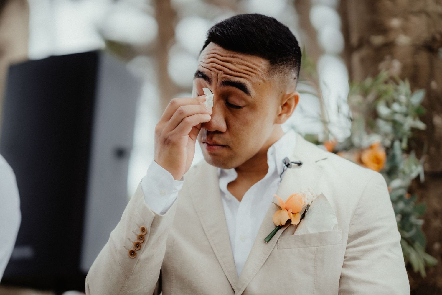 Emotional groom in a cream suit with a peach boutonniere wipes a tear from his eye during the wedding ceremony.