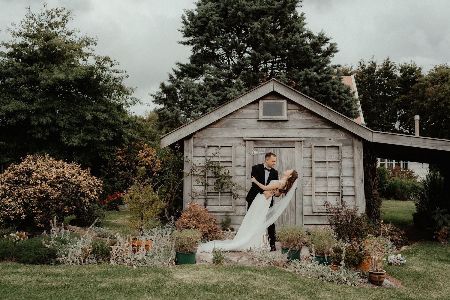 Groom dipping the bride in a romantic pose outside a rustic wooden cottage surrounded by garden plants.