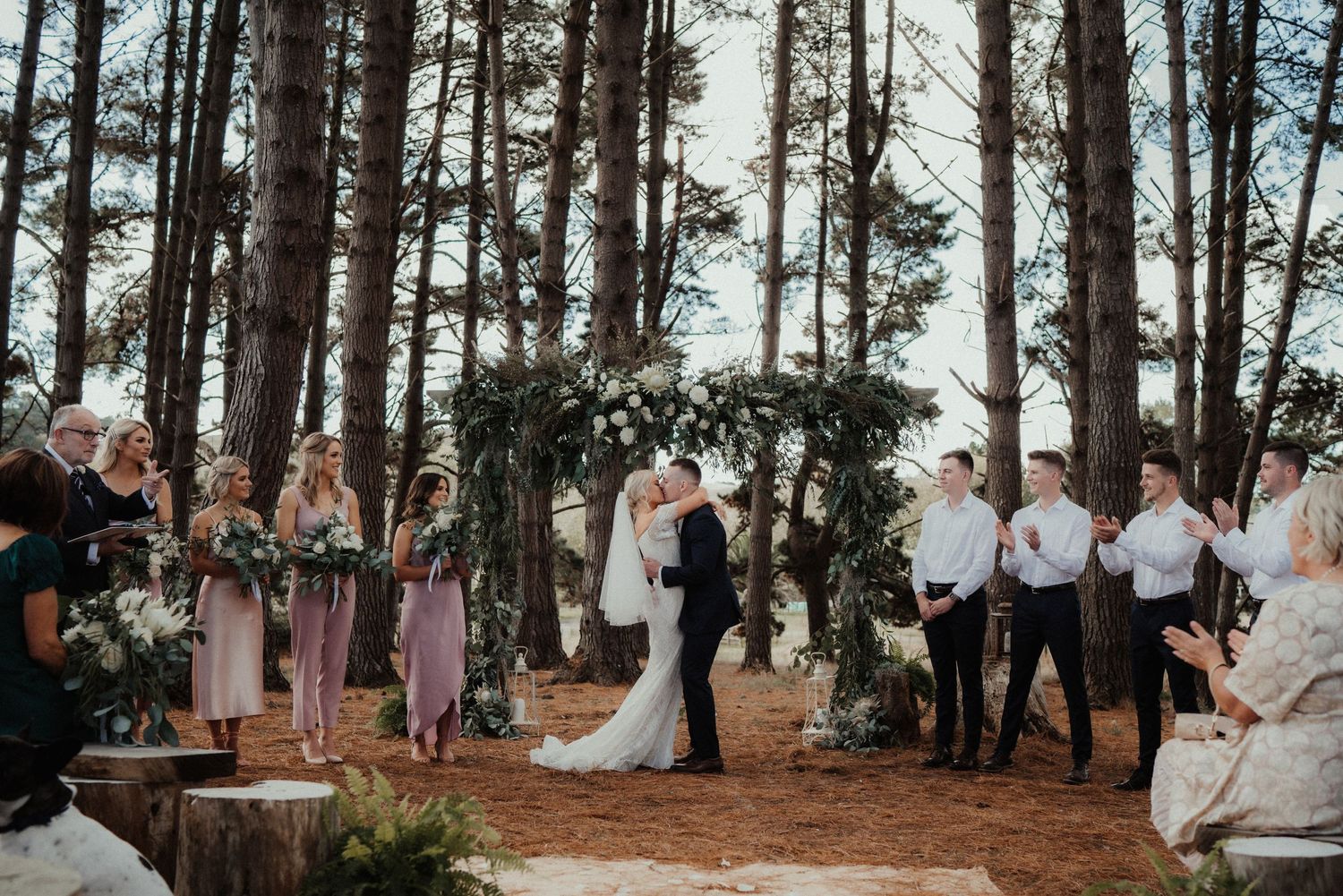 Bride and groom share a kiss under a greenery and white flower arch while bridesmaids, groomsmen and guests look on in a pine forest.