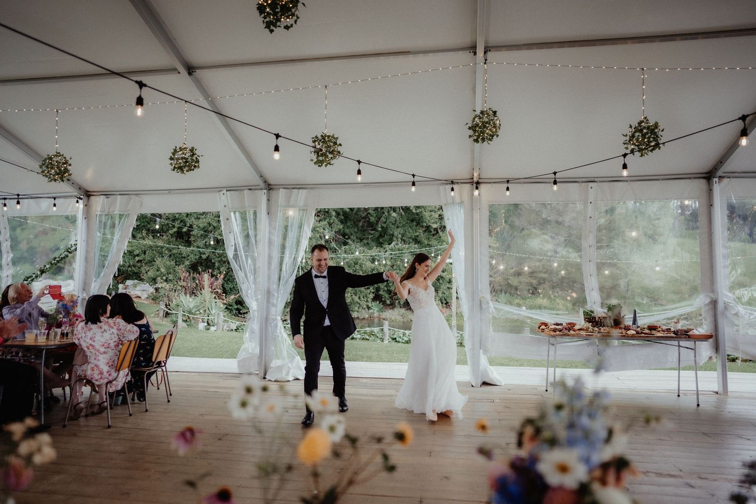 Bride and groom dance inside a white wedding marquee decorated with hanging foliage balls, string lights, and wildflower arrangements.