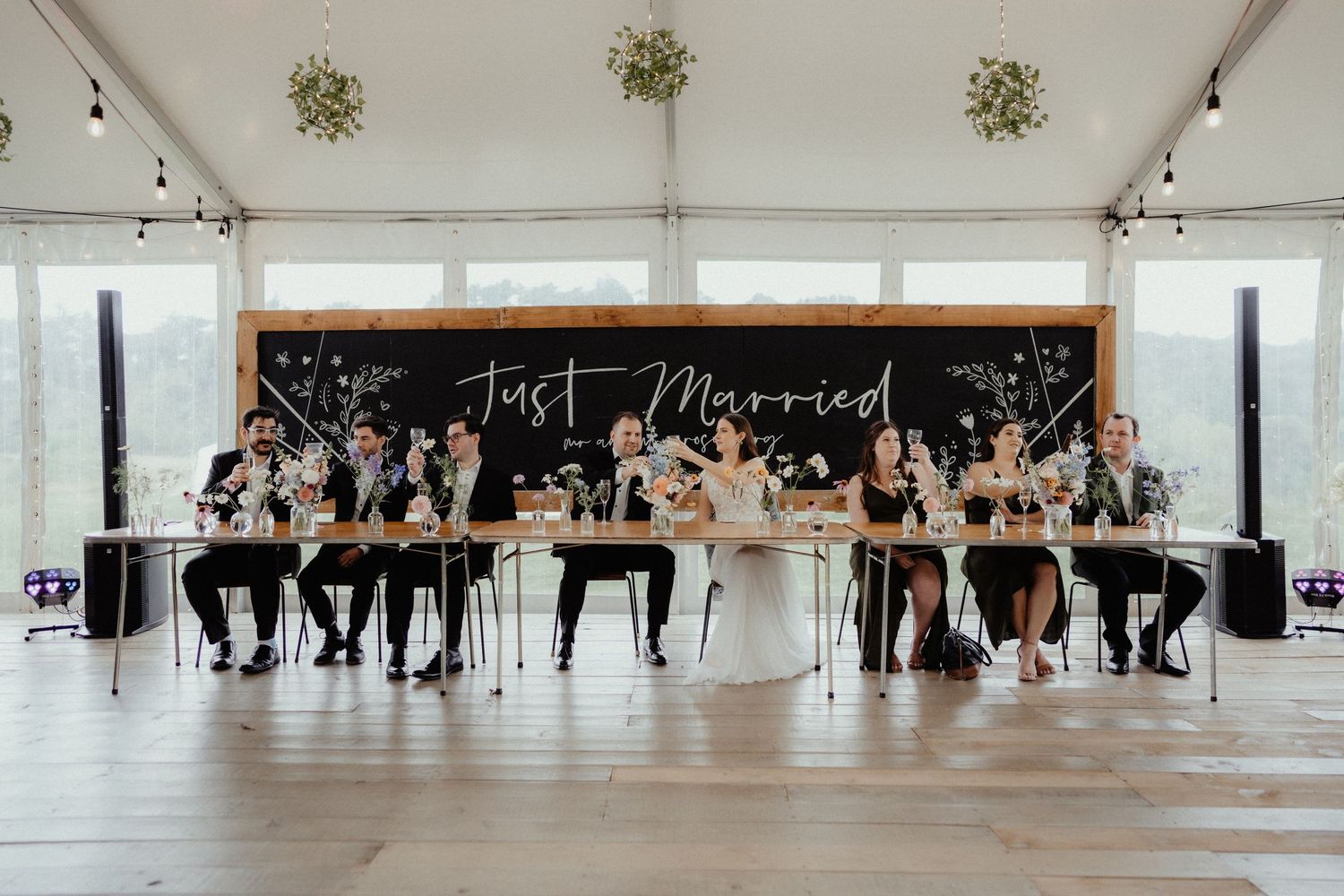 Bridal party sits at a long head table beneath a Just Married chalkboard sign inside a bright wedding marquee.