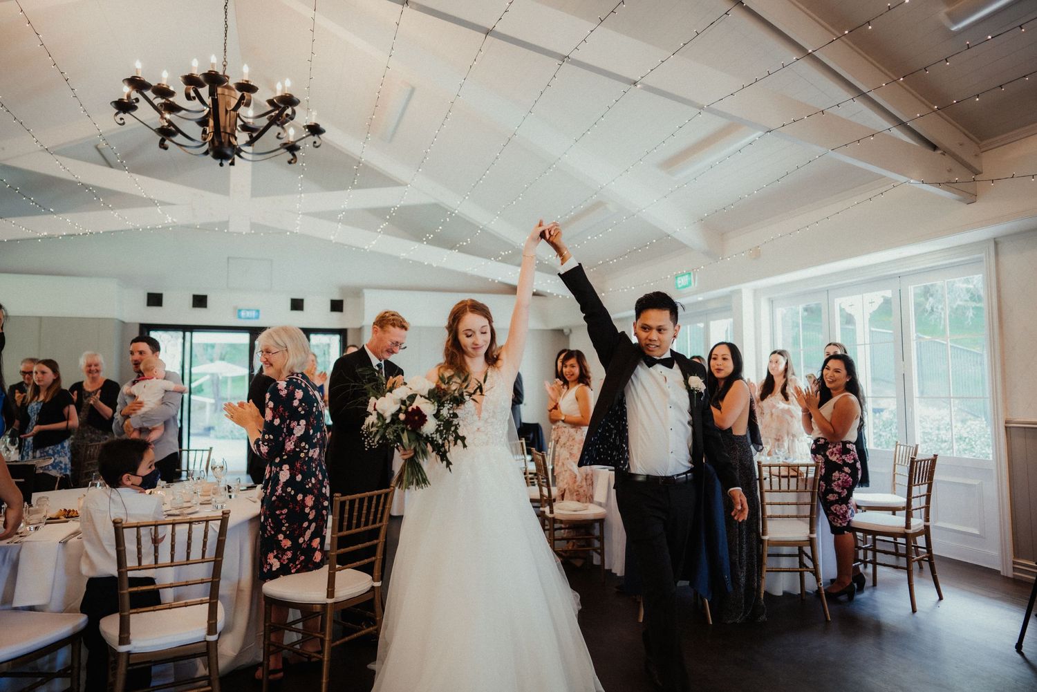 Bride and groom raise their joined hands as they walk down the aisle of a bright venue while smiling guests look on.