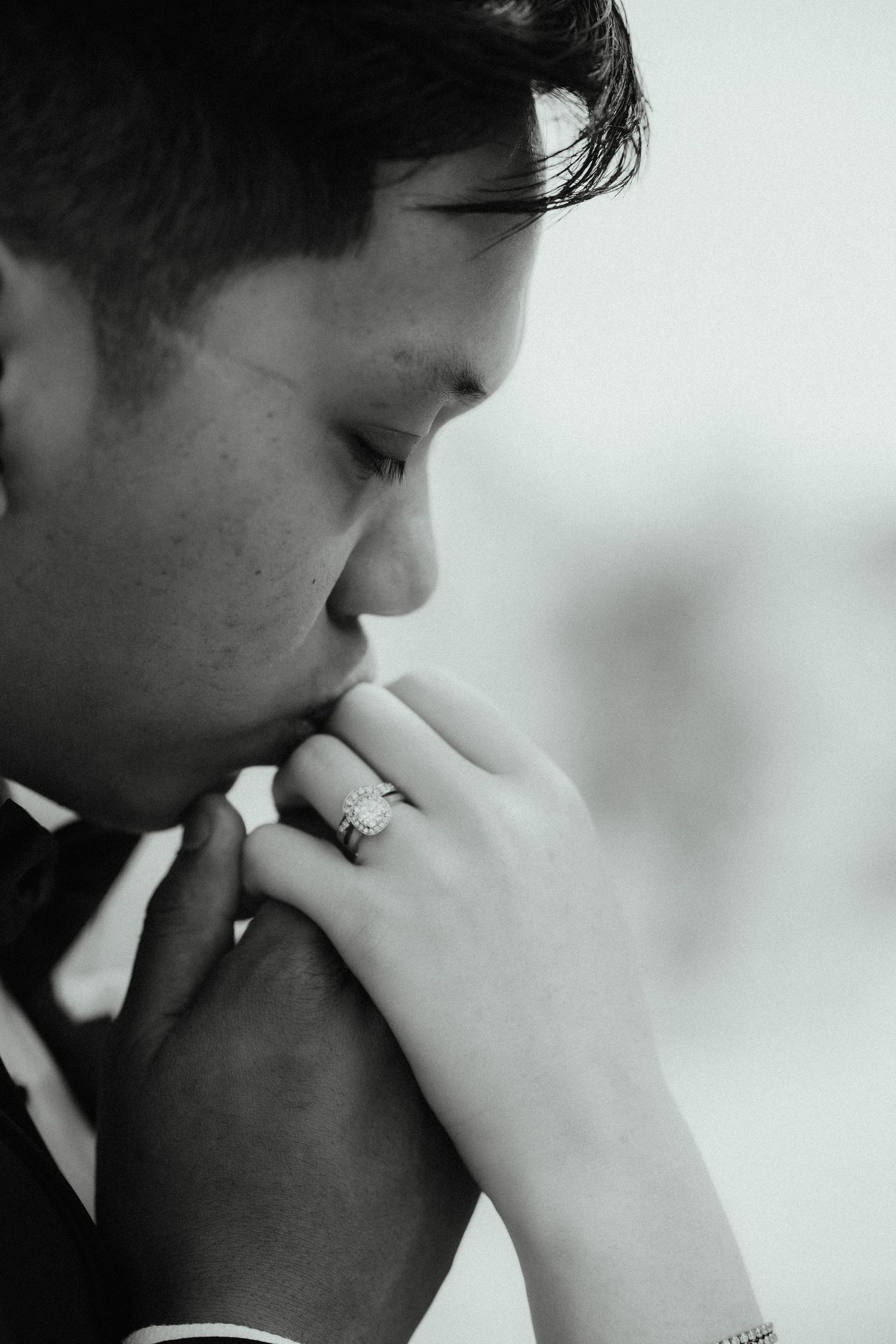 Black and white close-up of a groom kissing his partner's hand, showing her sparkling engagement ring.