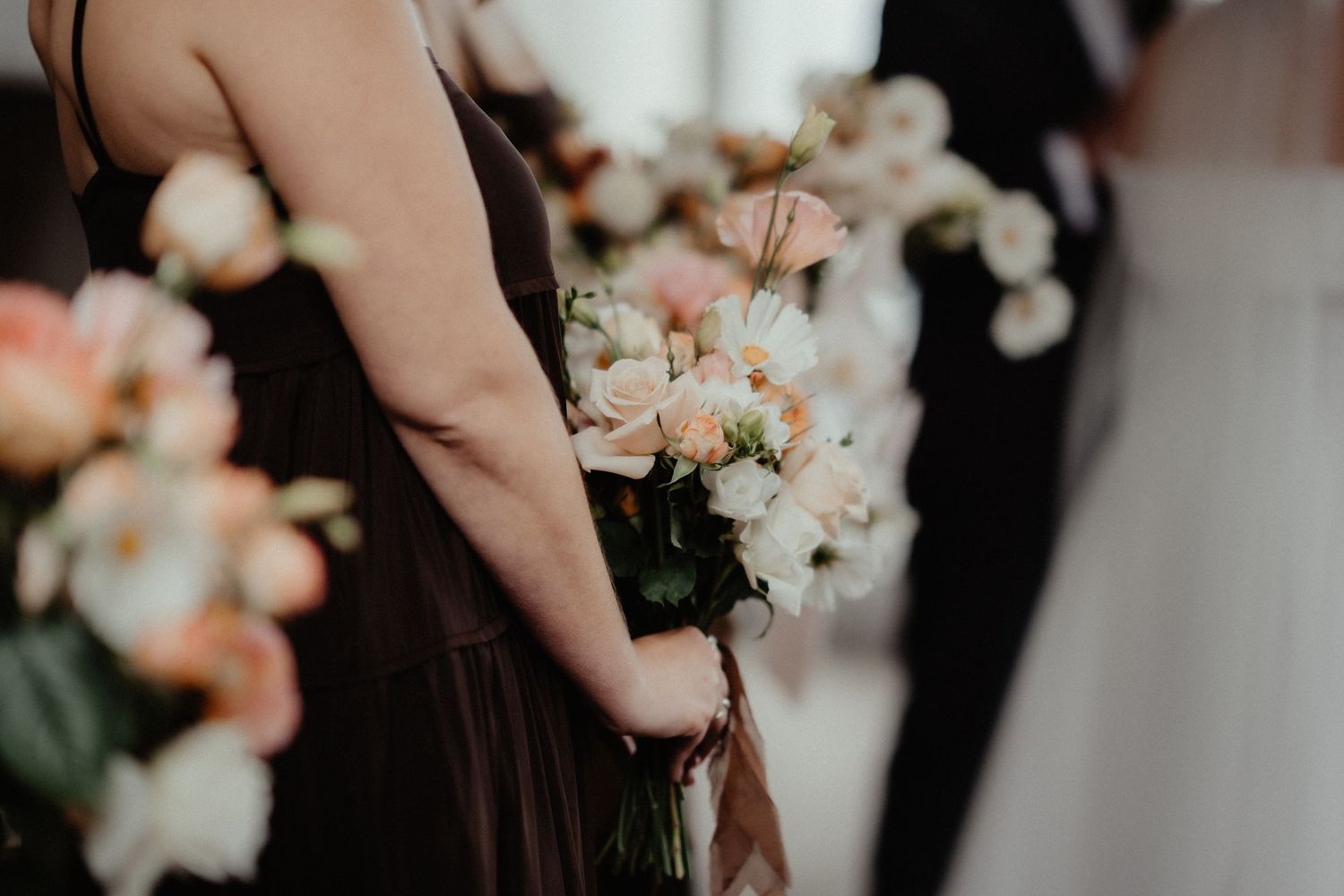 Bridesmaid in a dark brown dress holds a bouquet of peach and white flowers during a wedding ceremony.