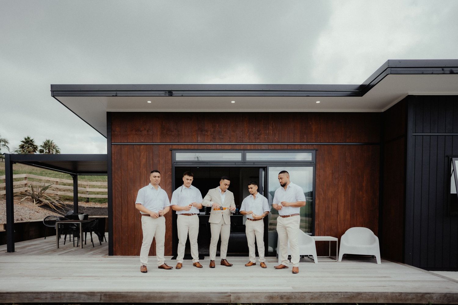 Five groomsmen in light linen outfits stand on a modern timber deck in front of a dark wood-clad house.