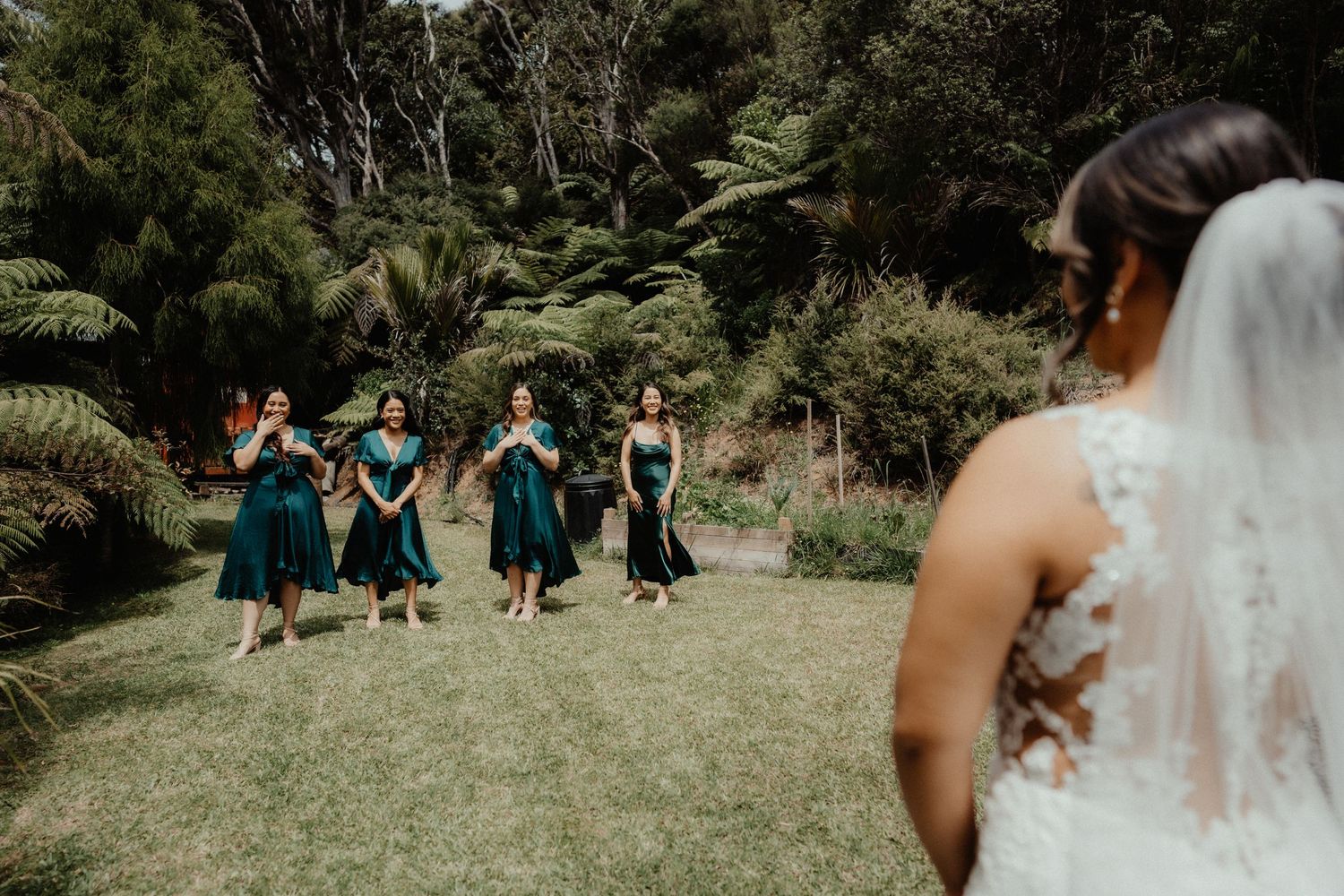 A bride in a lace gown and veil faces four bridesmaids in emerald green dresses standing on a lawn surrounded by ferns.