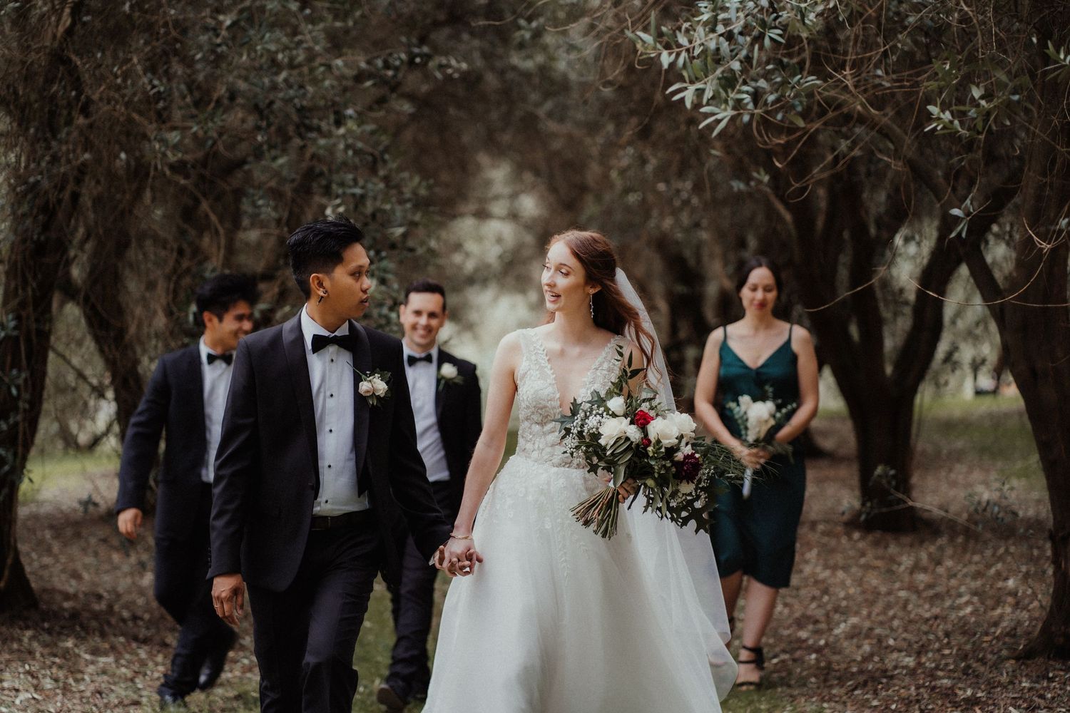 A smiling bride holding a rose bouquet walks hand in hand with her groom as the wedding party strolls through the olive trees.