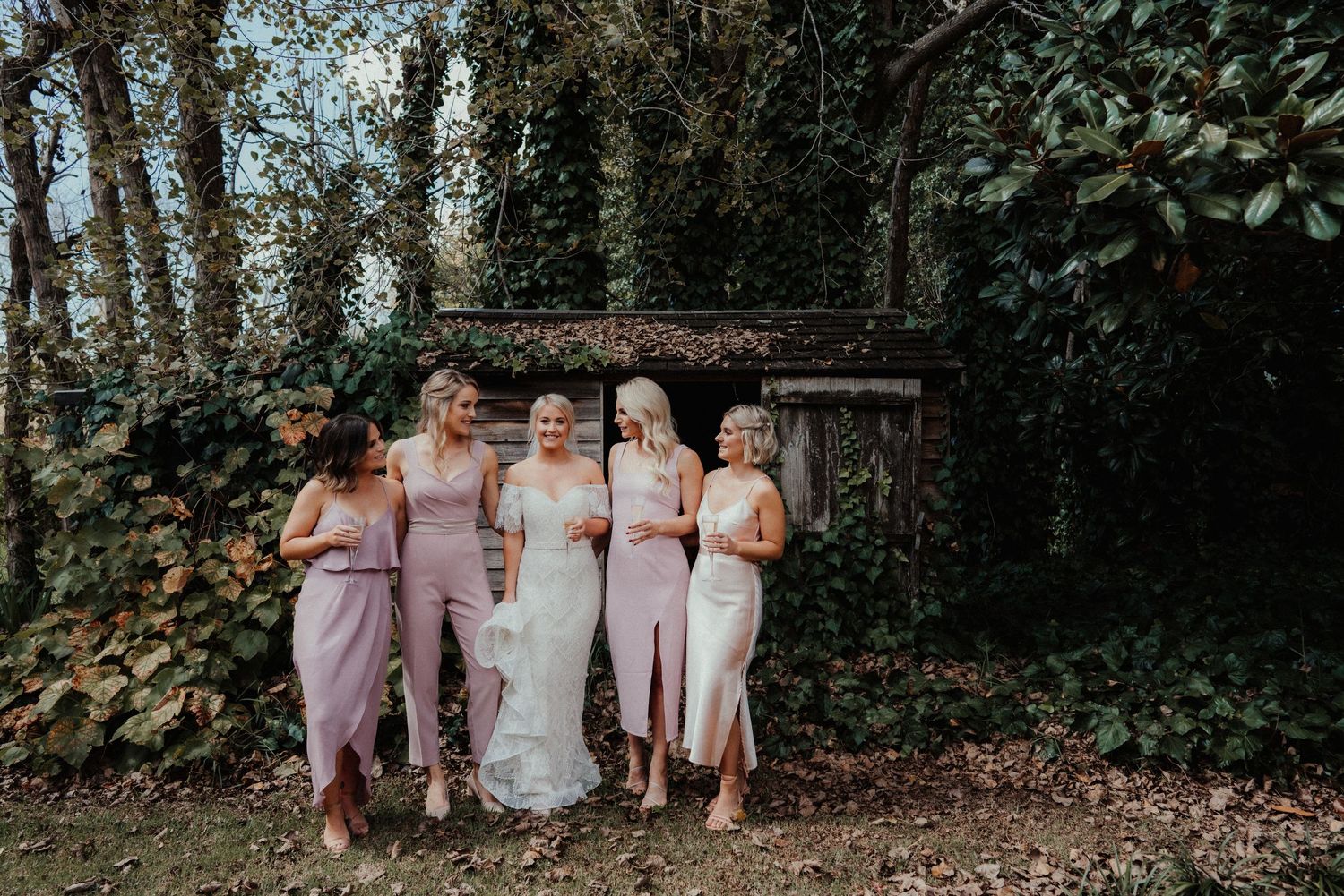 Bride and four bridesmaids in blush and ivory dresses laugh together beside an ivy-covered stone shed at a wedding.