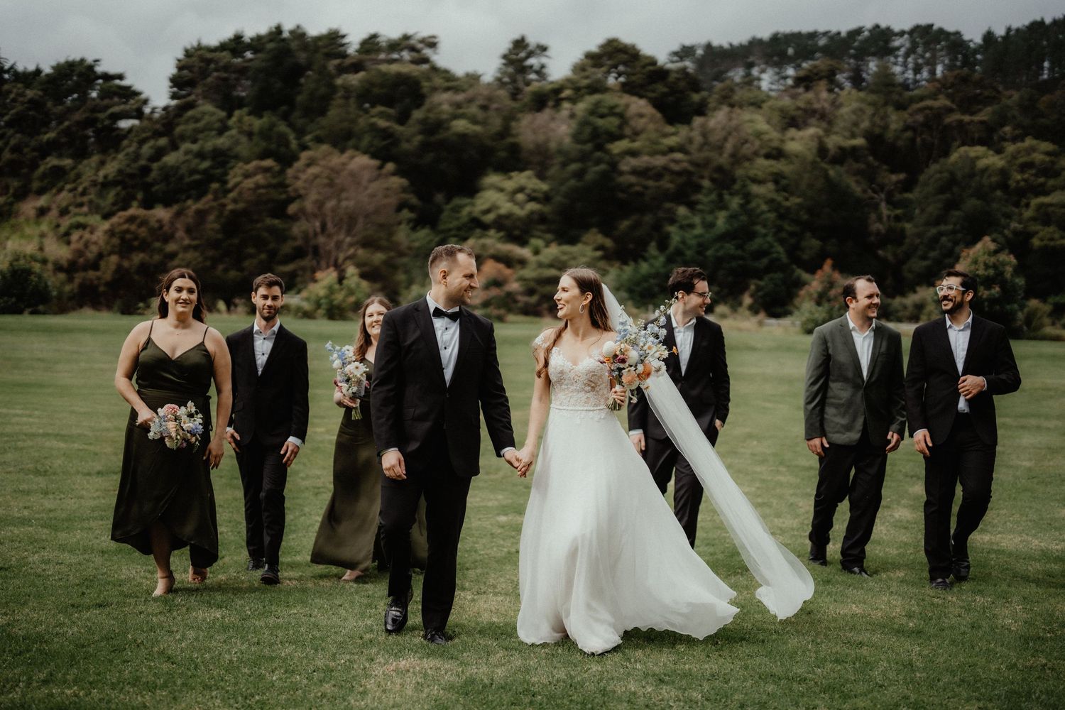 The bride and groom hold hands and walk across a lawn with smiling bridesmaids and groomsmen, her veil trailing behind.