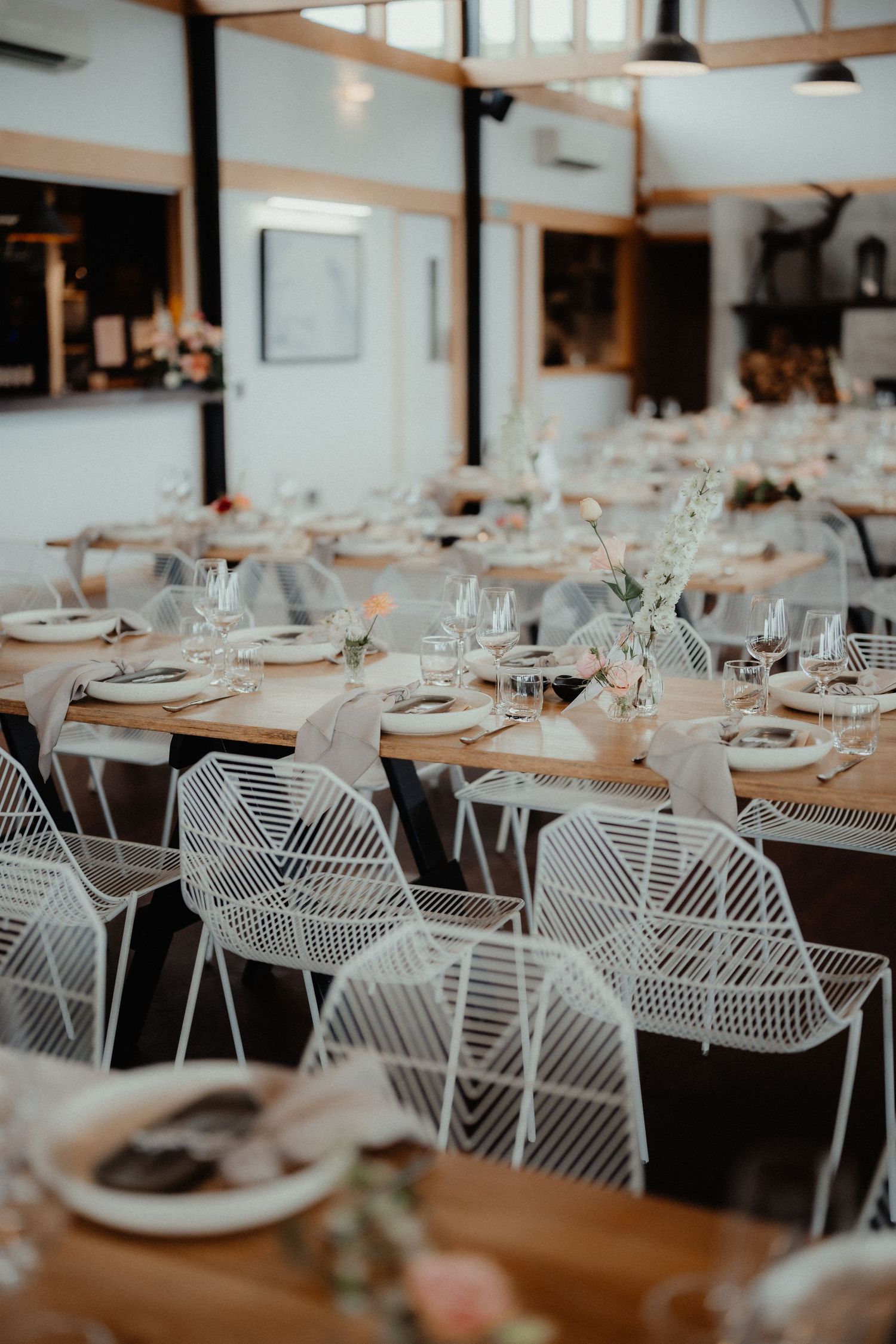 Elegant wedding reception tables with white wire chairs, place settings, napkins and fresh floral centerpieces in a bright venue.