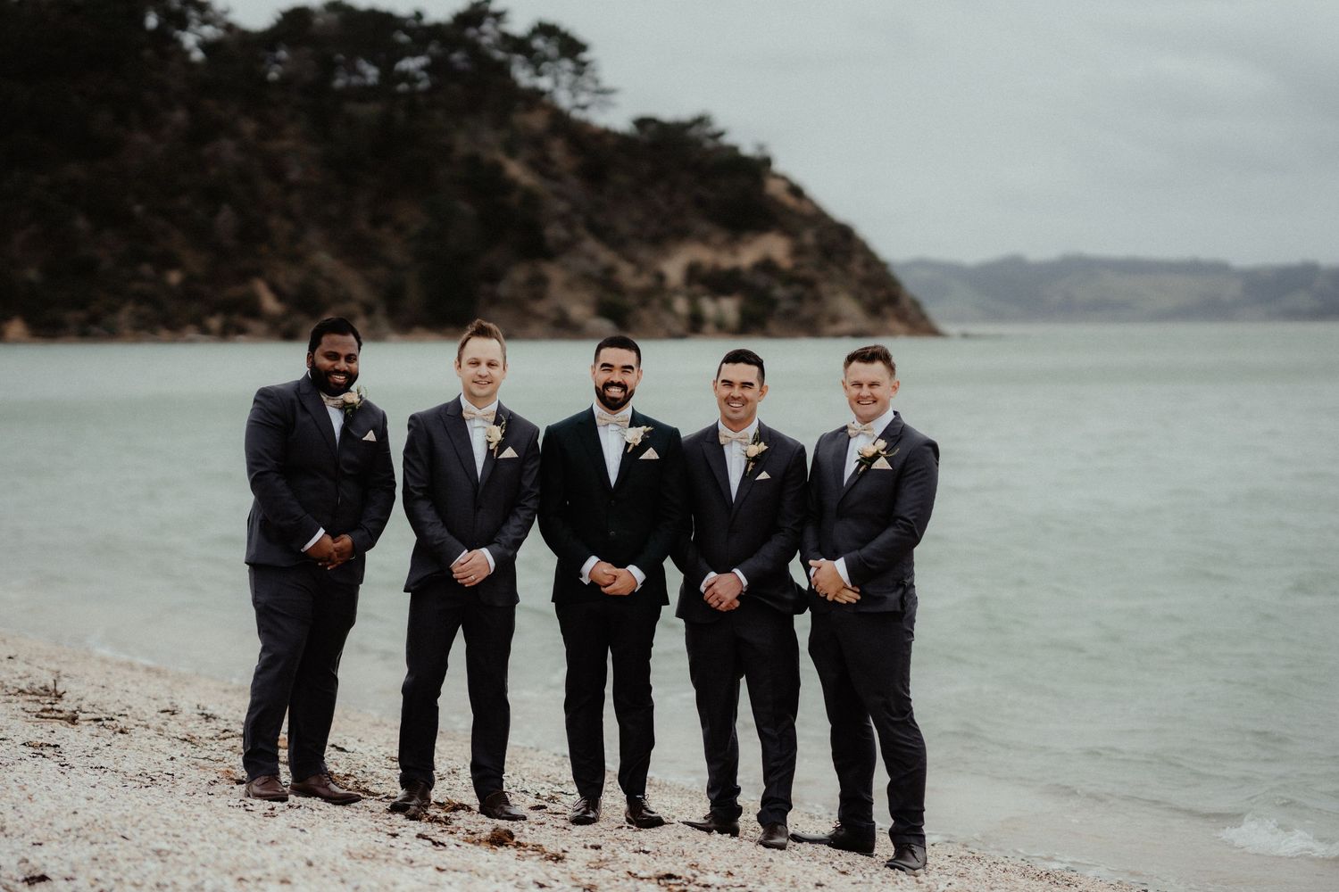 Five groomsmen in dark suits stand in a row on a sandy beach with calm water and a forested headland behind them.
