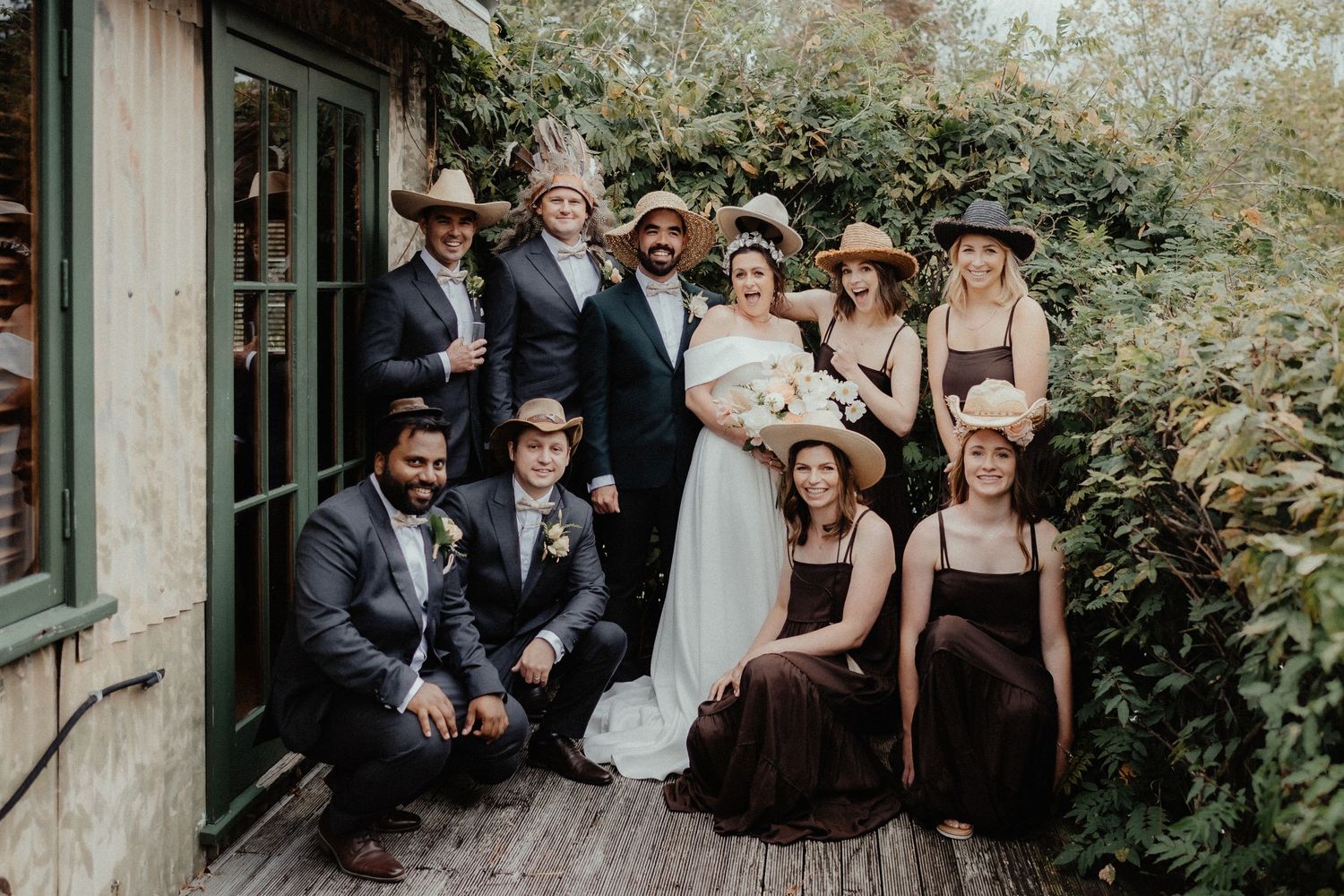 A bride and groom pose with their wedding party, all wearing cowboy hats and quirky headwear, on a wooden porch beside green foliage.