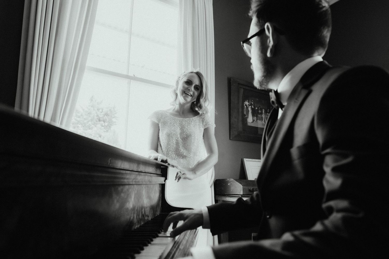 Black and white wedding photo of a groom in a bow tie playing a piano while his bride smiles beside a sunlit window.
