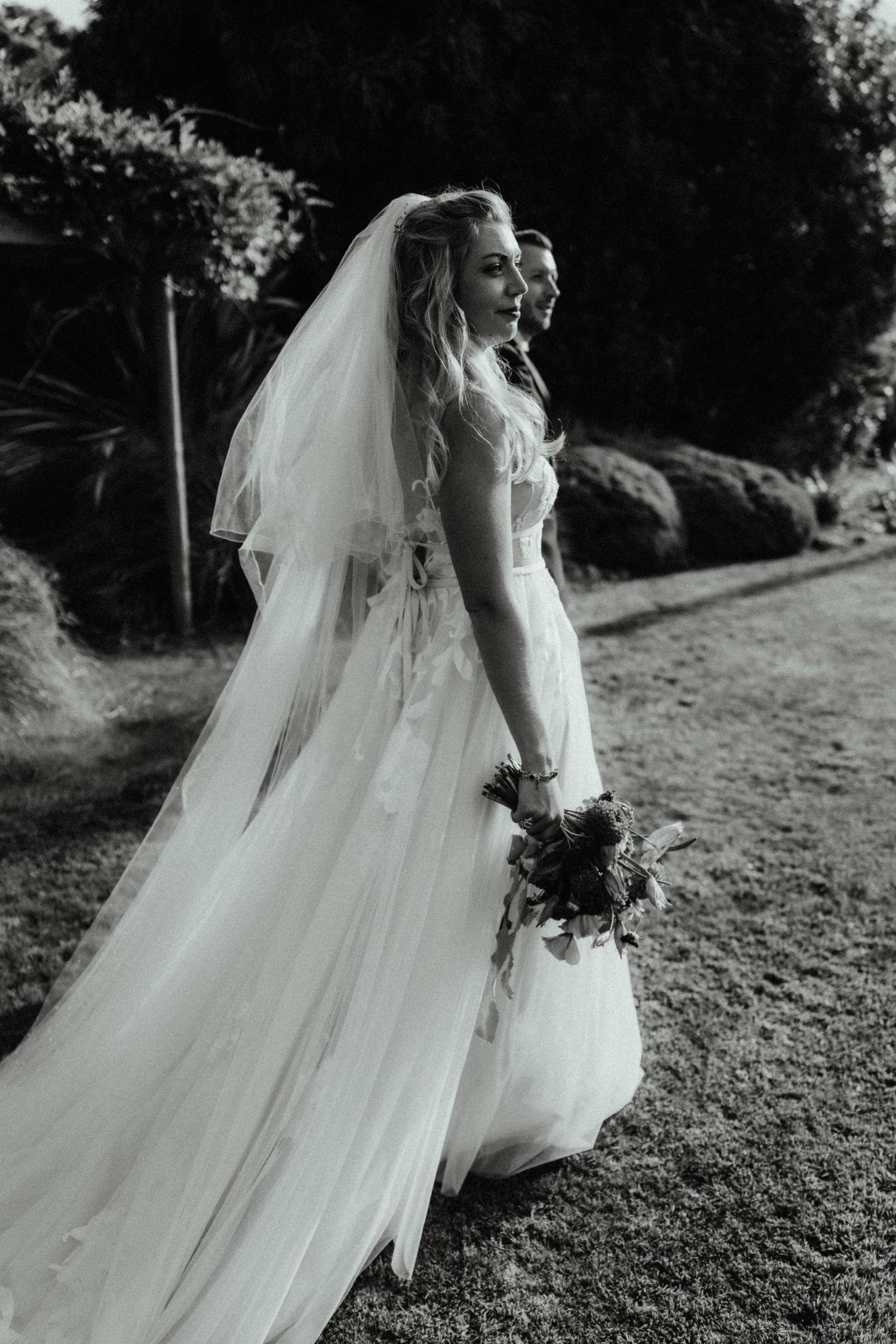 Black and white photo of a bride in a long tulle veil holding a bouquet as she looks over her shoulder in a garden.