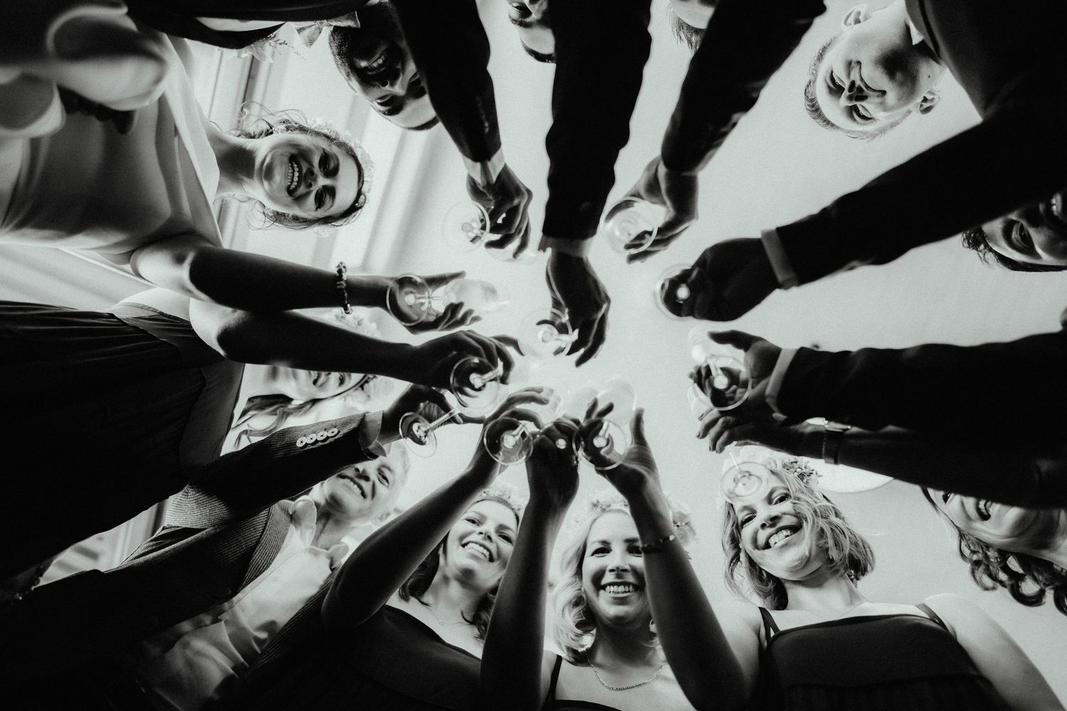 Black and white low-angle photo of smiling bridesmaids and groomsmen reaching hands together in a circle above the camera.