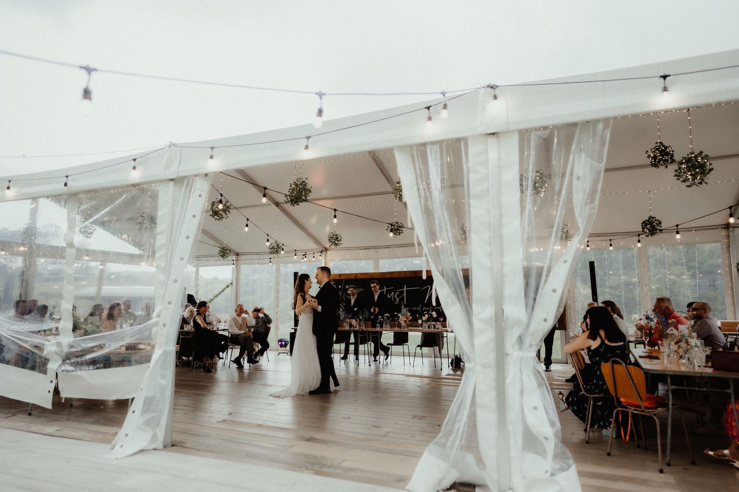 A bride and groom stand on the dance floor inside a clear marquee with draped curtains and festoon lights as guests watch from their tables.