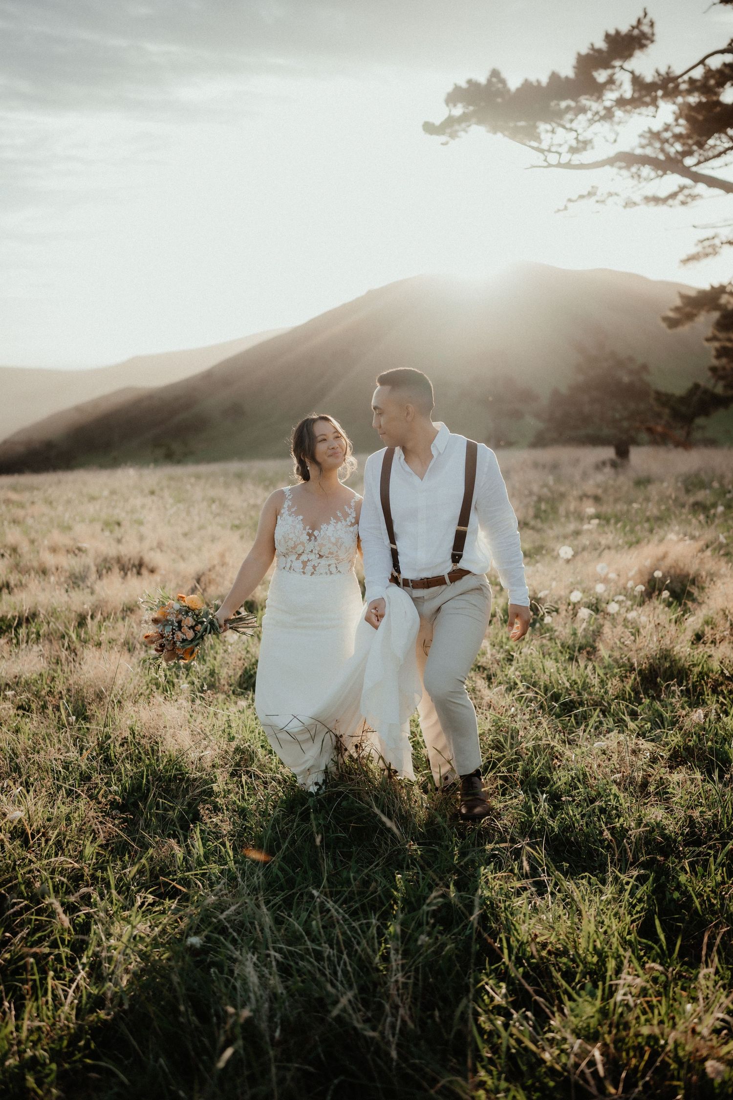 Smiling bride in a lace gown and groom in suspenders walk through a sunlit hillside meadow holding a colorful bouquet.