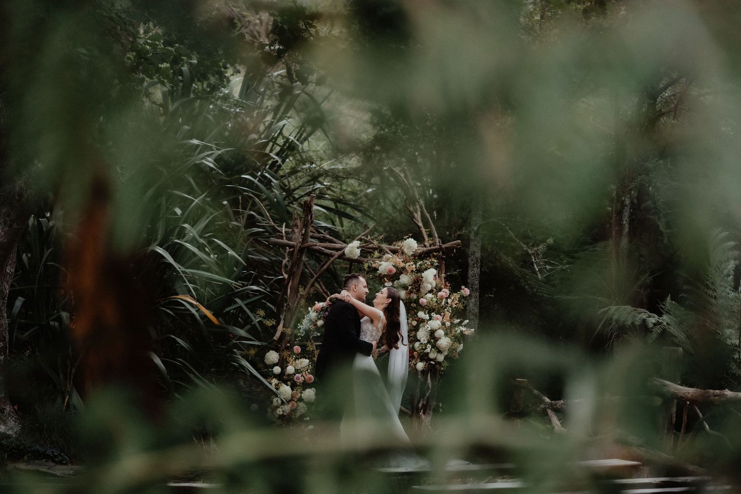 Bride and groom embrace in front of a floral wedding arch, framed by blurred green foliage in a forest ceremony setting.