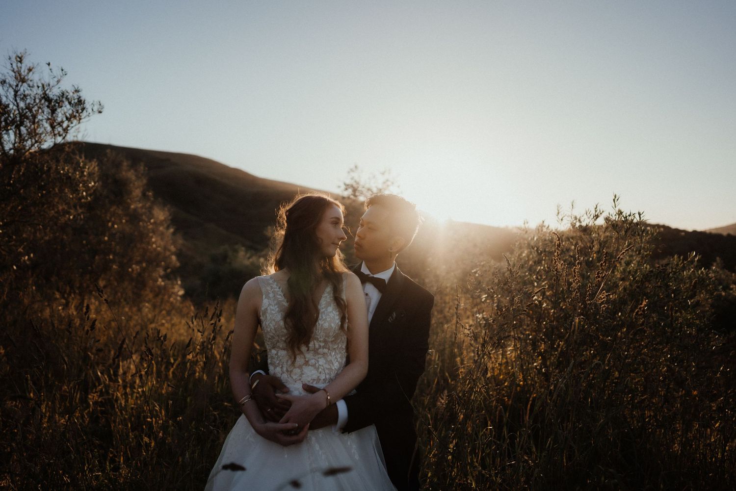 Bride and groom embrace in golden sunset light among autumn shrubs with rolling hills behind them.