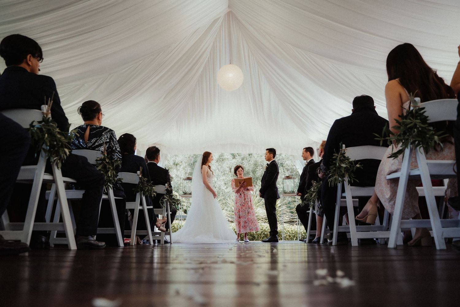 Wedding guests sit on white chairs as a bride and groom exchange vows under a draped white tent with a hanging globe light.