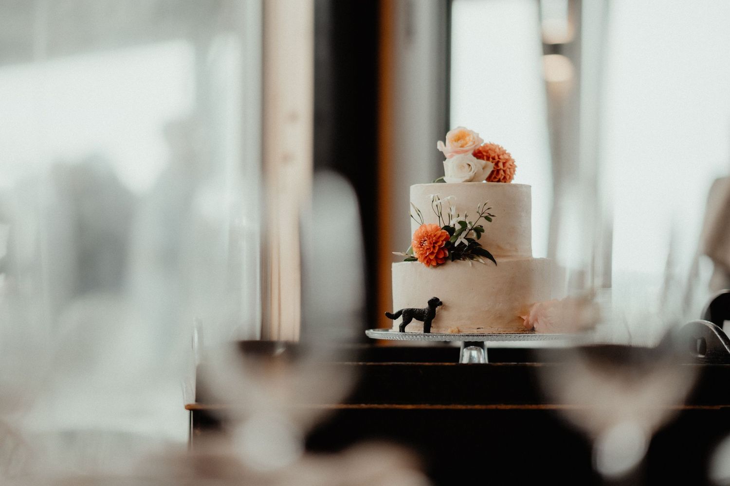 Two-tier wedding cake topped with peach and white flowers sits on a table beside a small black dog figurine.