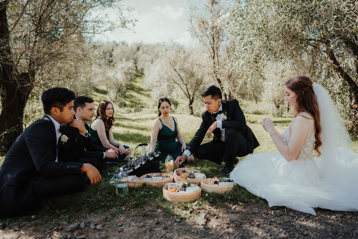A bride, groom and wedding guests share a casual picnic on the ground beneath olive trees at an outdoor wedding.