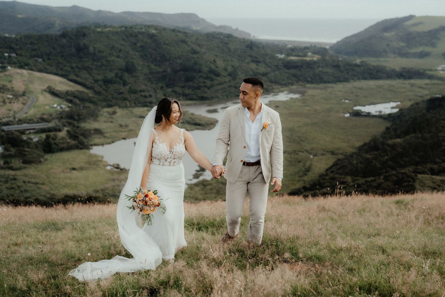 Bride and groom hold hands and walk through a grassy hilltop with a bouquet, overlooking a river valley.