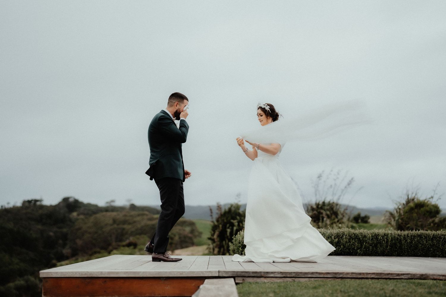 Groom in a dark suit wipes his eyes during an emotional first look as the bride in a long veil stands on a wooden platform.