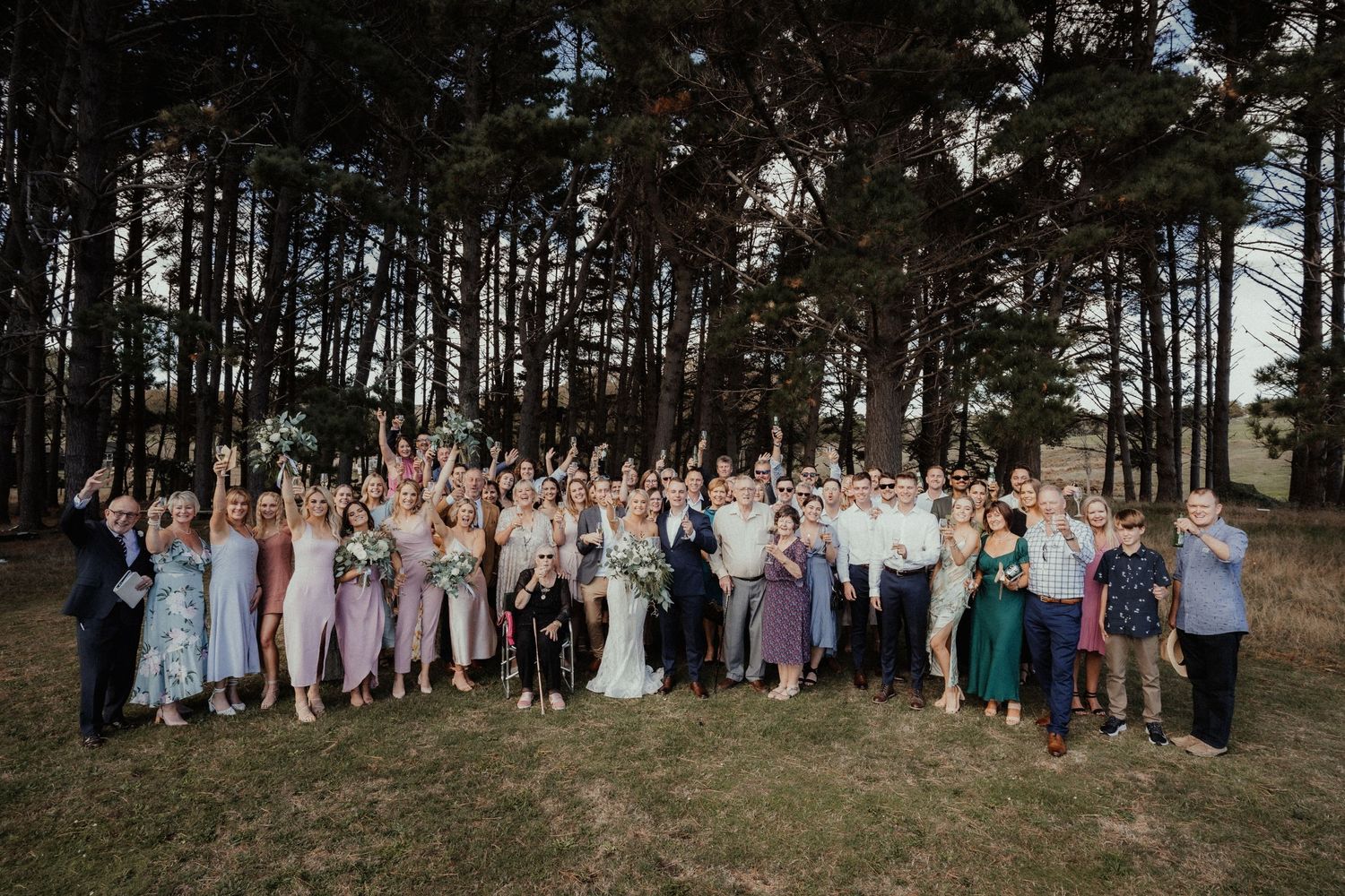 Groom stands in the center of a row of nine groomsmen in white shirts and sunglasses posing on a grassy field under a blue sky.