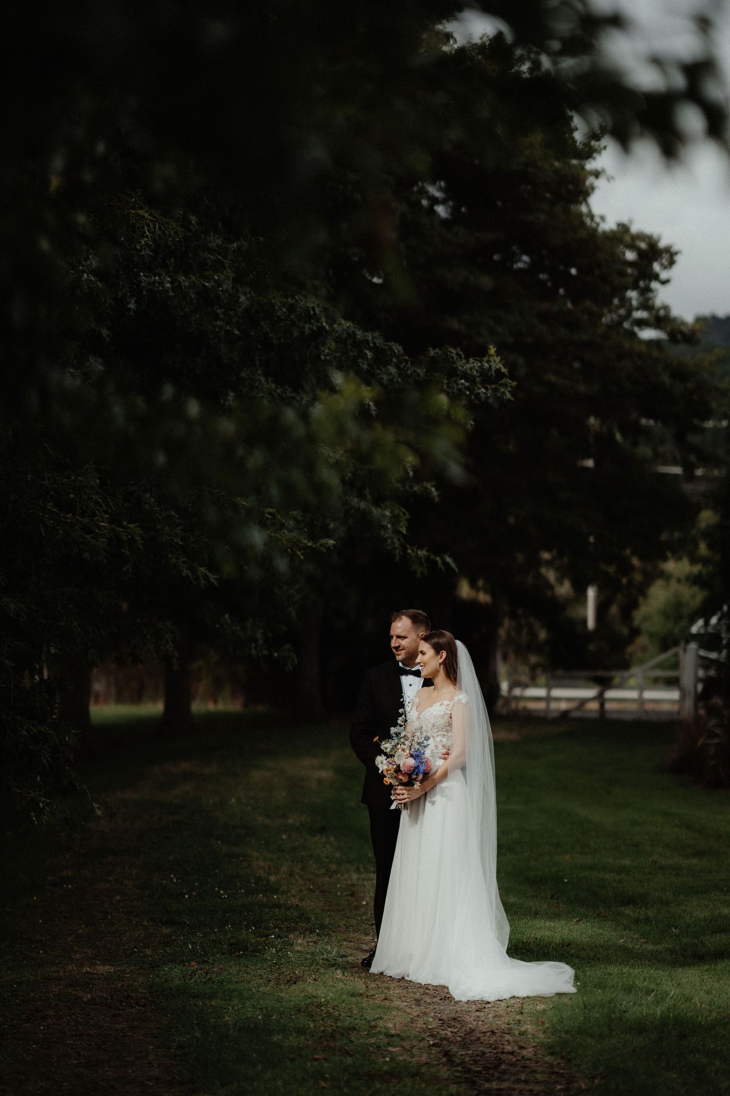 Bride in a long veil and groom in a black suit stand together on a grassy path beneath tall dark trees.