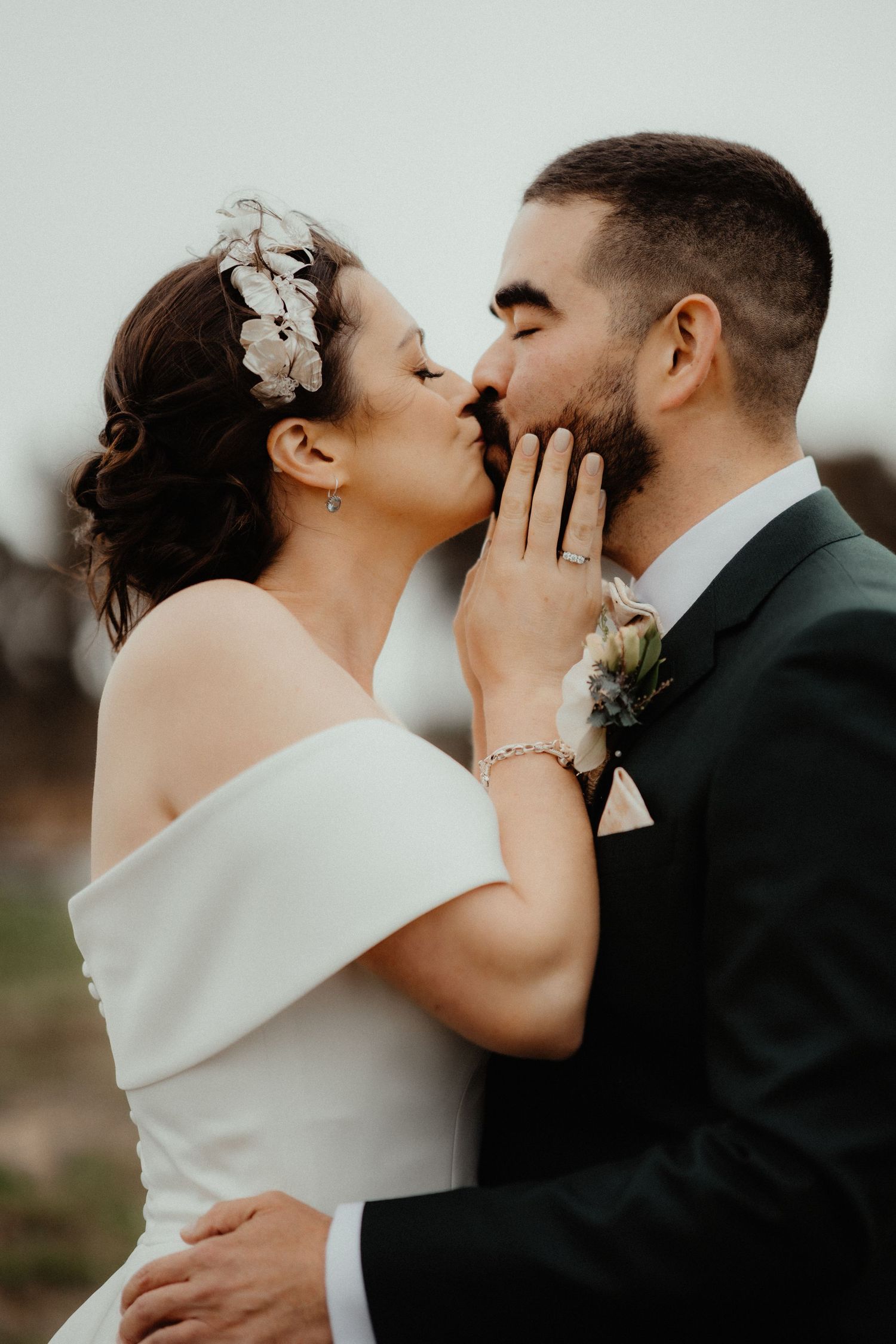 Bride in an off-shoulder white gown and floral headpiece kisses her groom in a dark suit with a boutonniere.