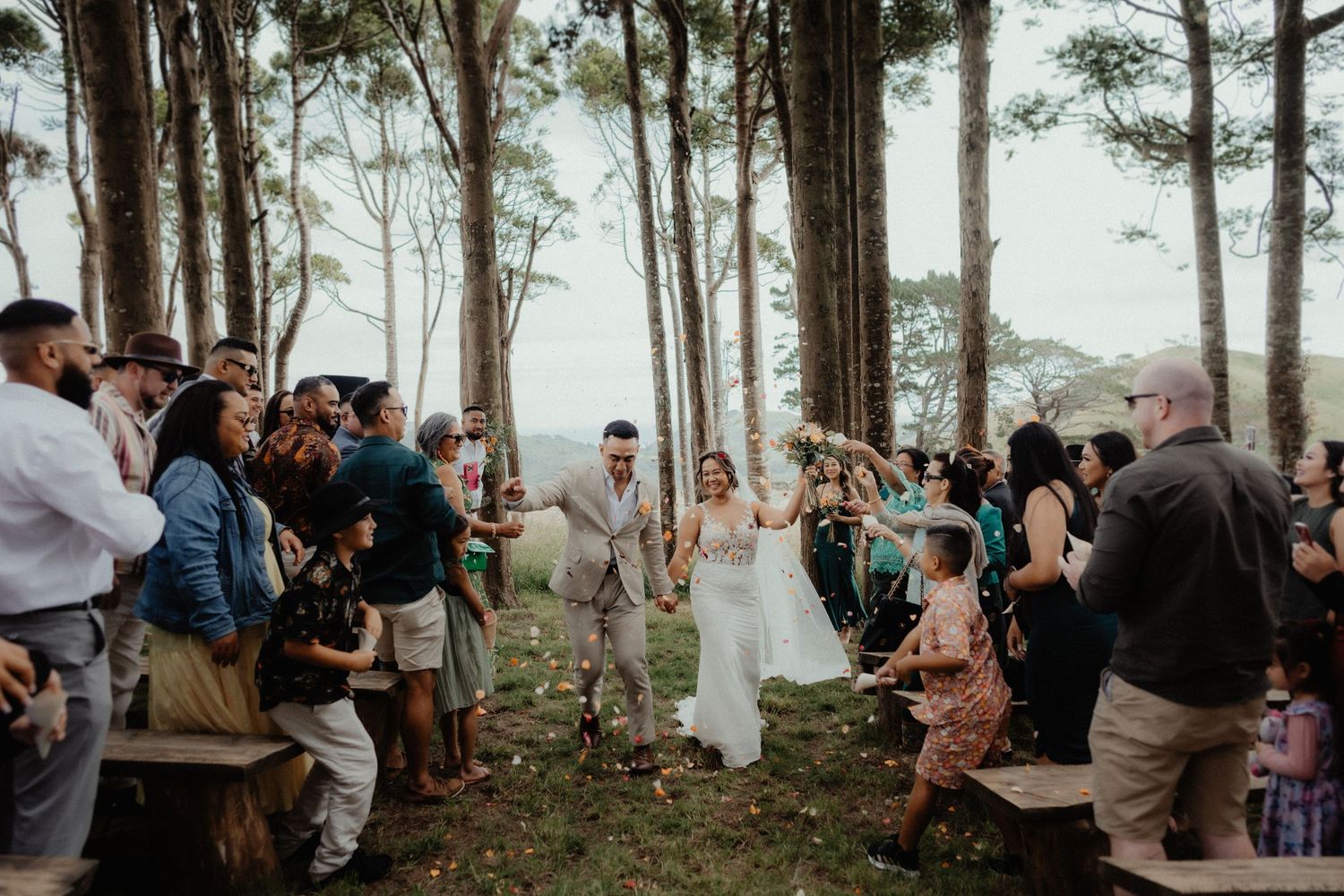 Bride and groom hold hands and walk down the aisle between cheering guests during an outdoor wedding ceremony in a forest.