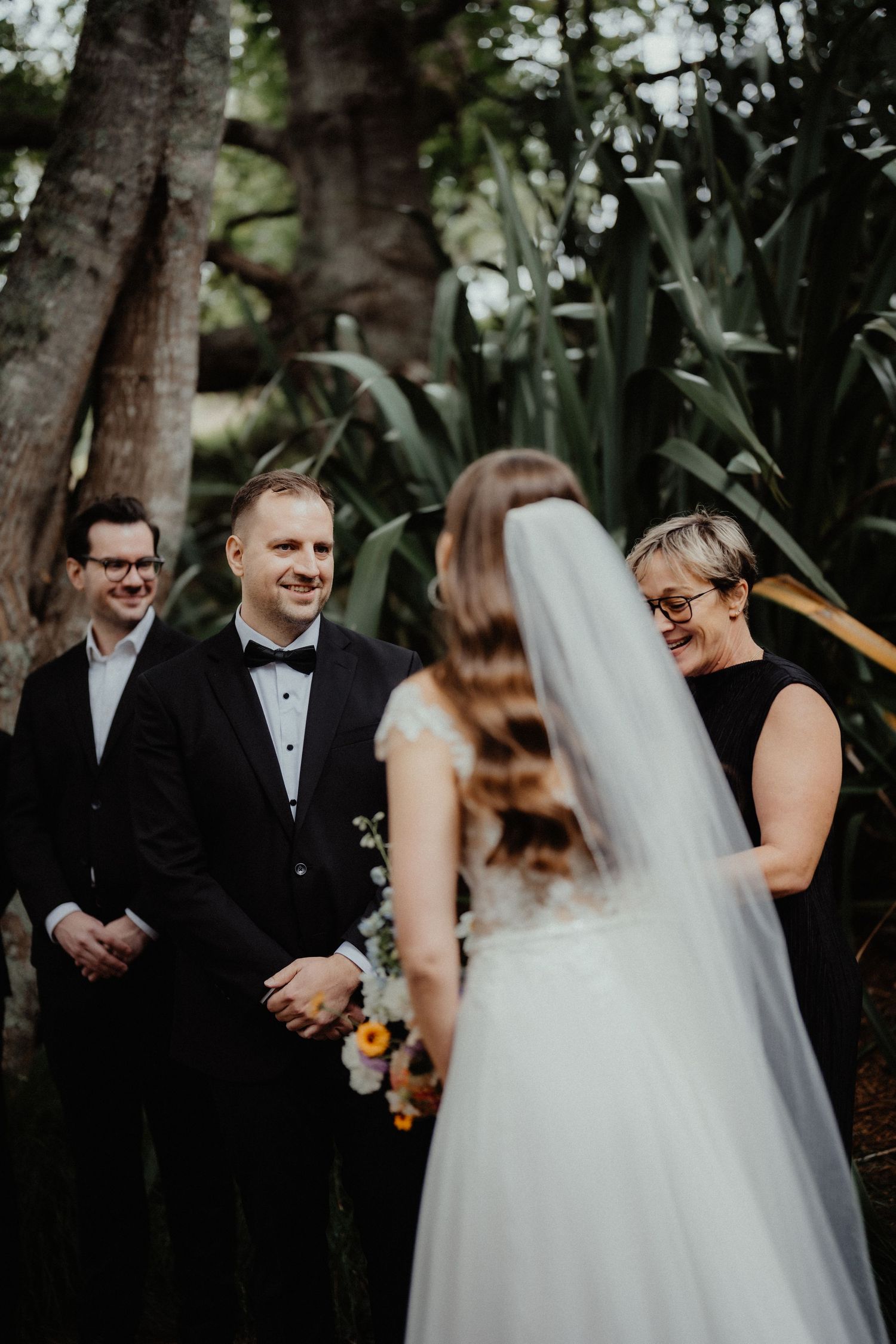 A bride in a lace gown and long veil faces her smiling groom in a bow tie as the celebrant and a groomsman look on outdoors.