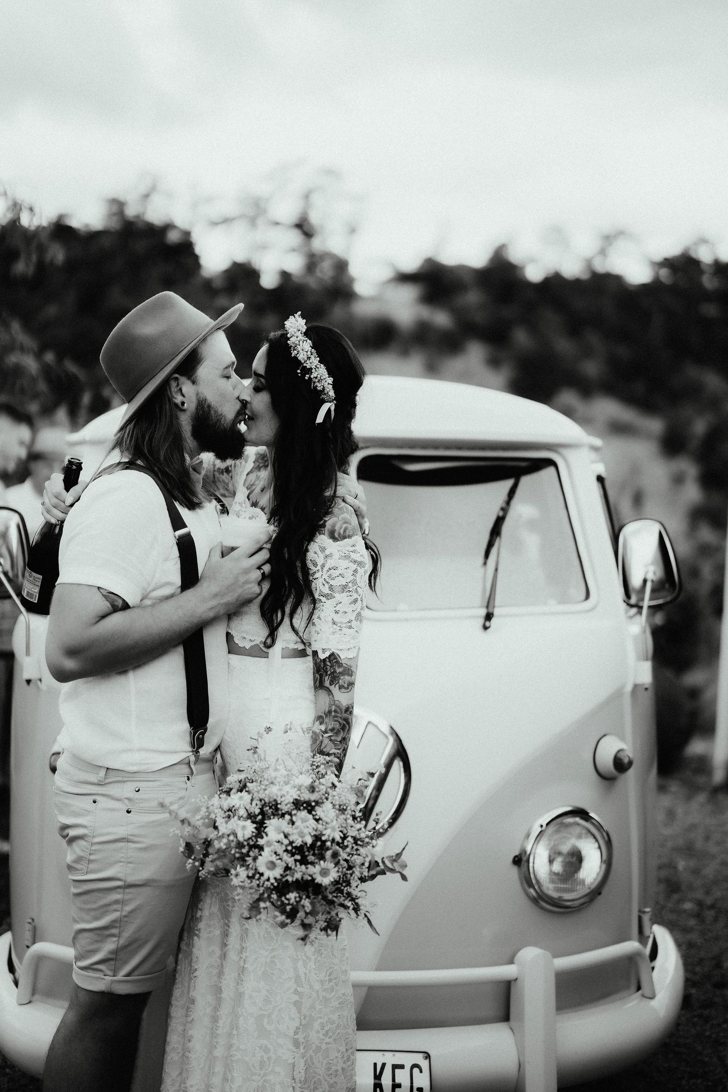 Black and white photo of a boho bride with a flower crown and groom in a hat kissing in front of a vintage camper van.